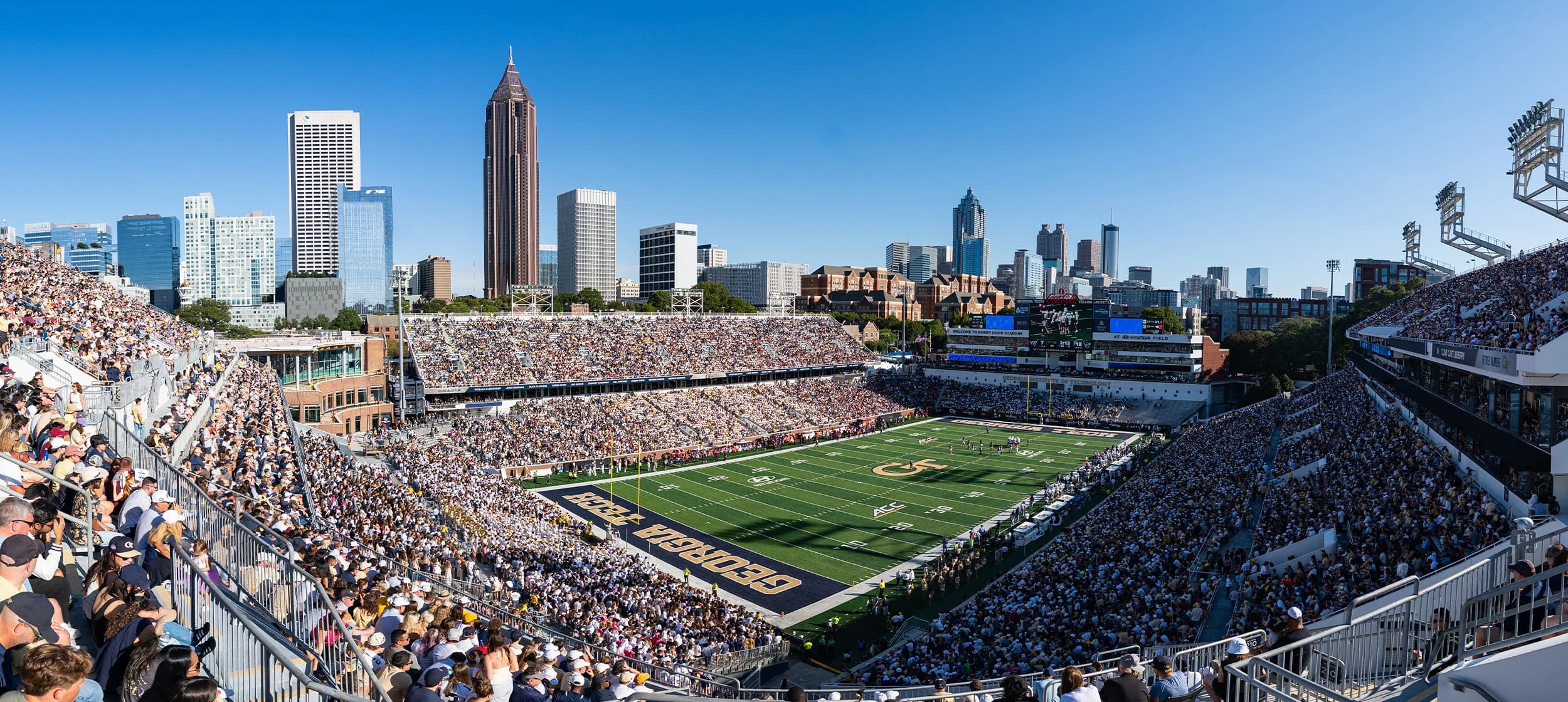 Bobby Dodd Stadium on a clear game-day afternoon — a packed bowl in white and gold, the GEORGIA TECH end zone below, and the Midtown Atlanta skyline rising directly beyond the stands