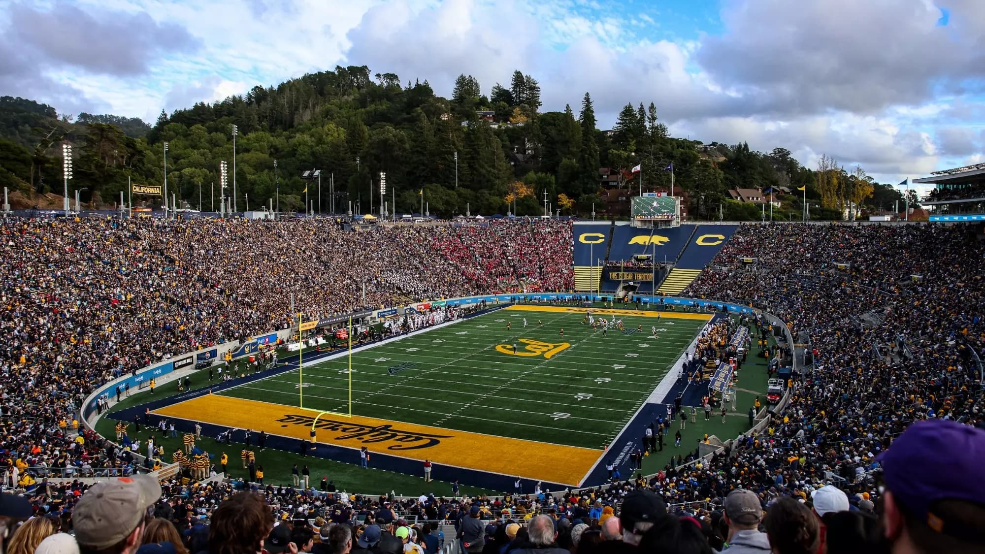California Memorial Stadium sold out on a fall afternoon — gold CALIFORNIA script end zones, the THIS IS BEAR TERRITORY banner across the south stands, and Tightwad Hill's wooded slope rising over the east rim under big Bay Area clouds