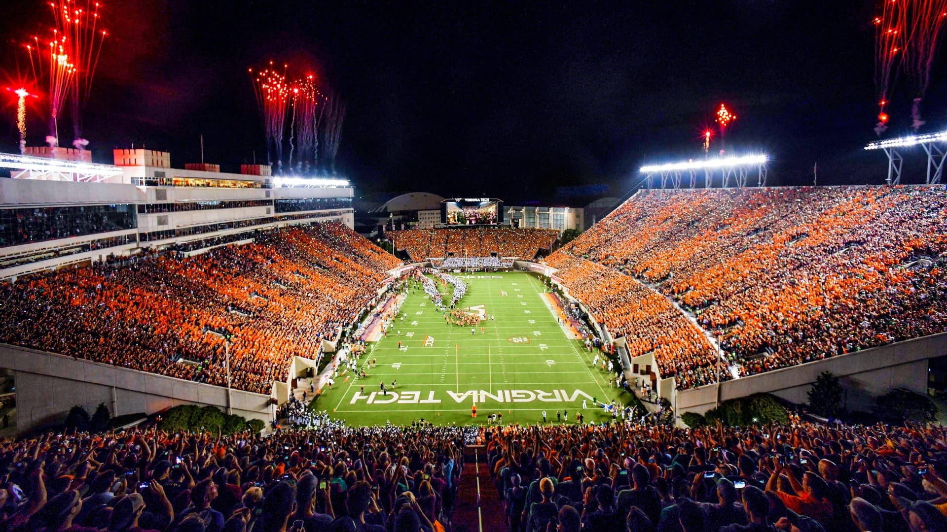 Lane Stadium at night from the south end zone — a sold-out orange-effect crowd filling both decks, fireworks bursting over the rim lights, and the team streaming onto the field past the VIRGINIA TECH end zone