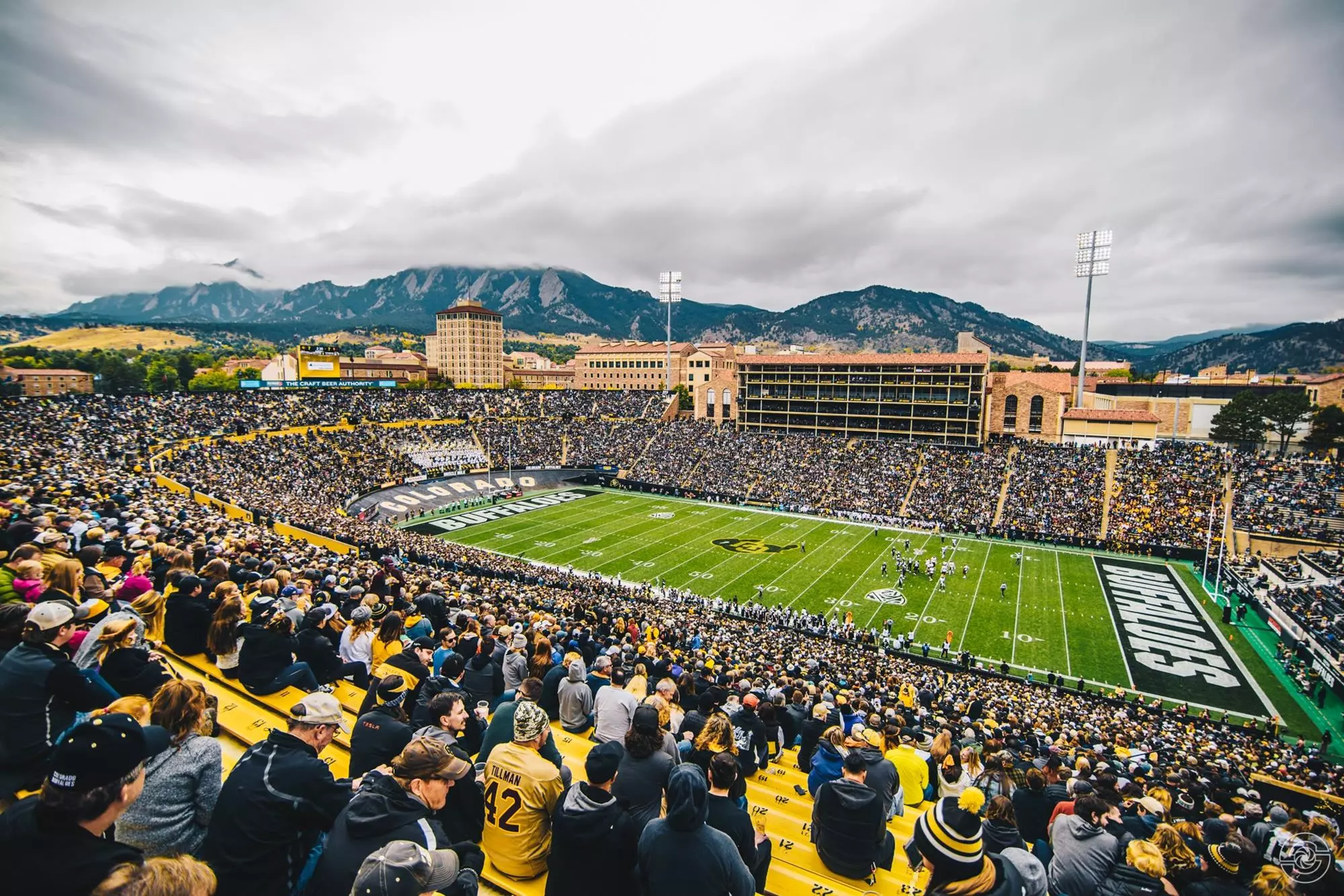 Folsom Field on game day — a sellout crowd on gold bleachers, COLORADO and BUFFALOES end zones framing the green, and the Flatirons rising into storm clouds behind the sandstone campus