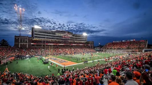 Gies Memorial Stadium at dusk — fireworks rising over the press box, an orange-and-blue crowd, and the ILLINOIS end zone below
