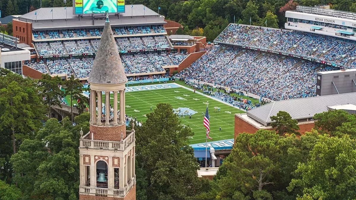 Kenan Stadium packed in Carolina Blue on a game Saturday, seen from above the pines — the Morehead-Patterson Bell Tower rising in the foreground and HEELS painted across the end zone