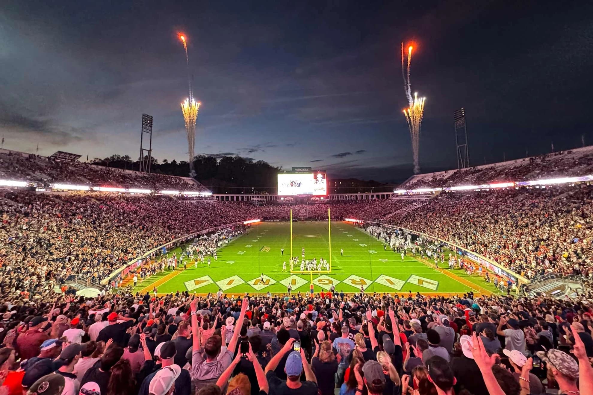 Scott Stadium at dusk on a sold-out night — fireworks climbing over both grandstands, the VIRGINIA diamond-pattern end zone glowing under the lights, and the crowd on its feet with arms raised