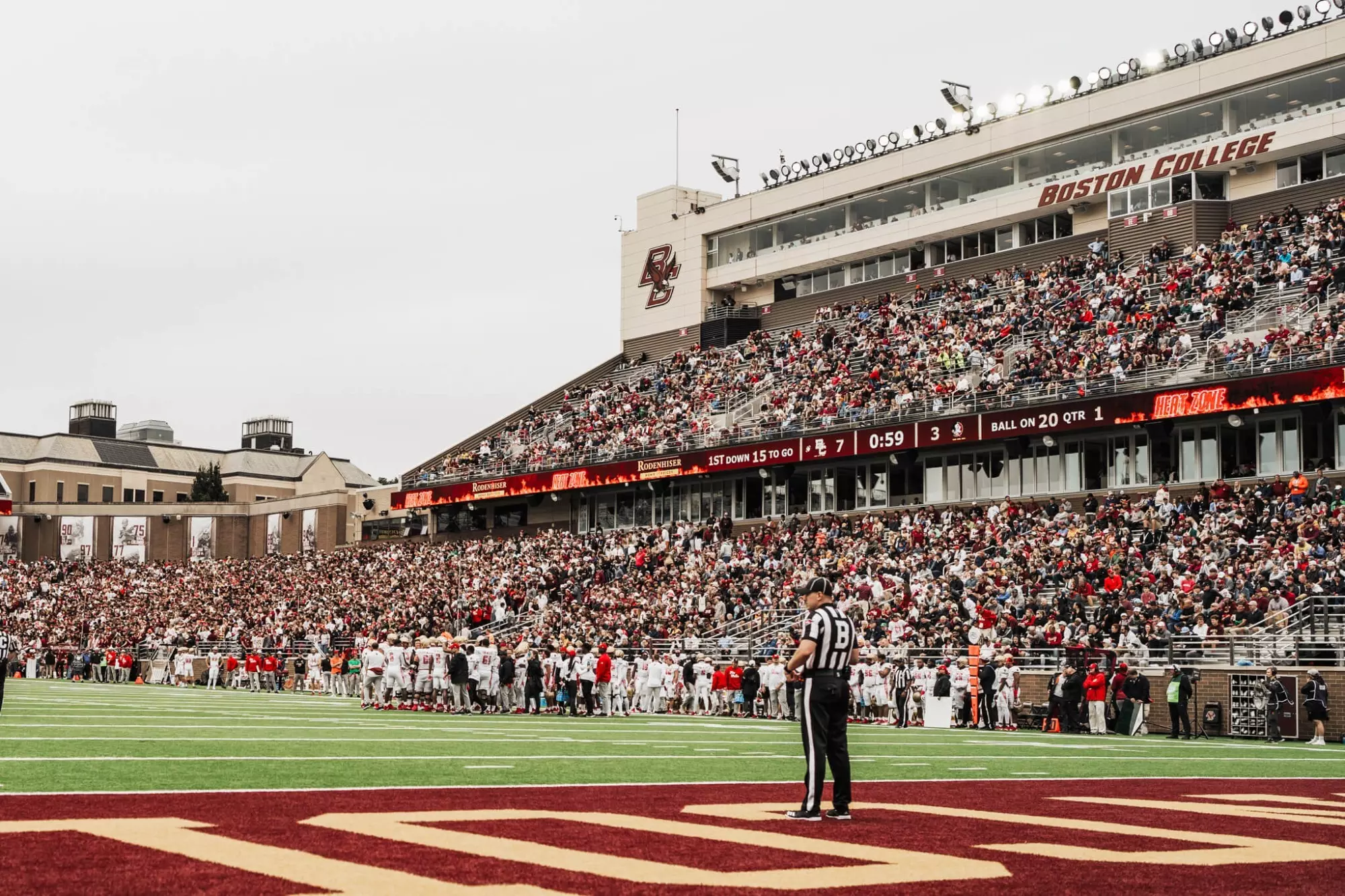 Field level inside a packed Alumni Stadium — the maroon end zone's gold letters up close, the crowd banked behind the BC sideline, and the Boston College press box rising over the Heights