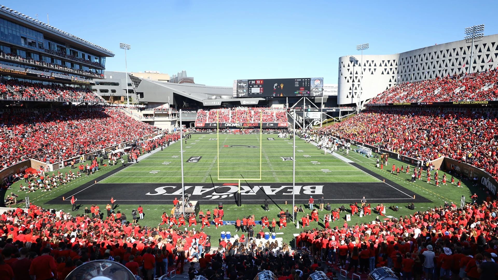 Nippert Stadium on a sold-out Saturday — the sunken bowl packed in red, the black BEARCATS end zone under the goalposts, and UC's glass-and-lattice campus buildings rising over every rim of the stadium