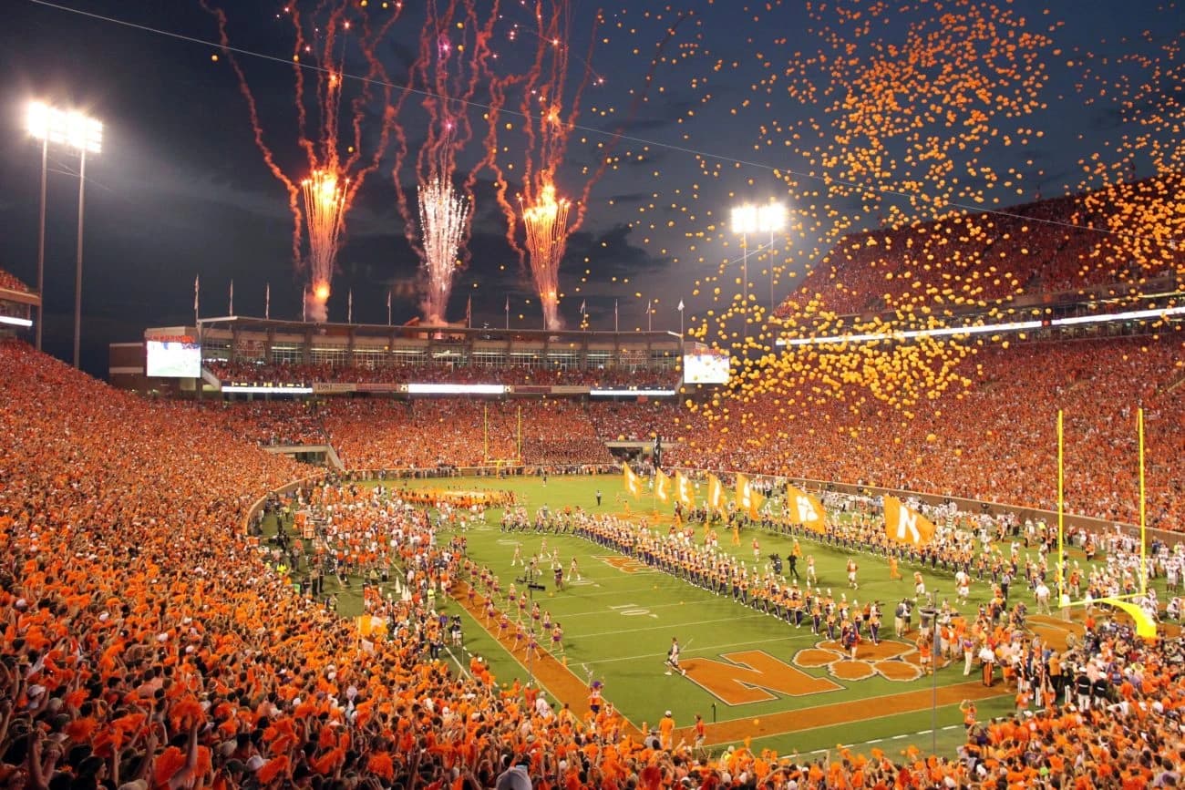 Memorial Stadium at night — fireworks over the press box, tens of thousands of orange balloons rising from a packed Death Valley, and C-L-E-M-S-O-N flags leading the team across the tiger-paw field