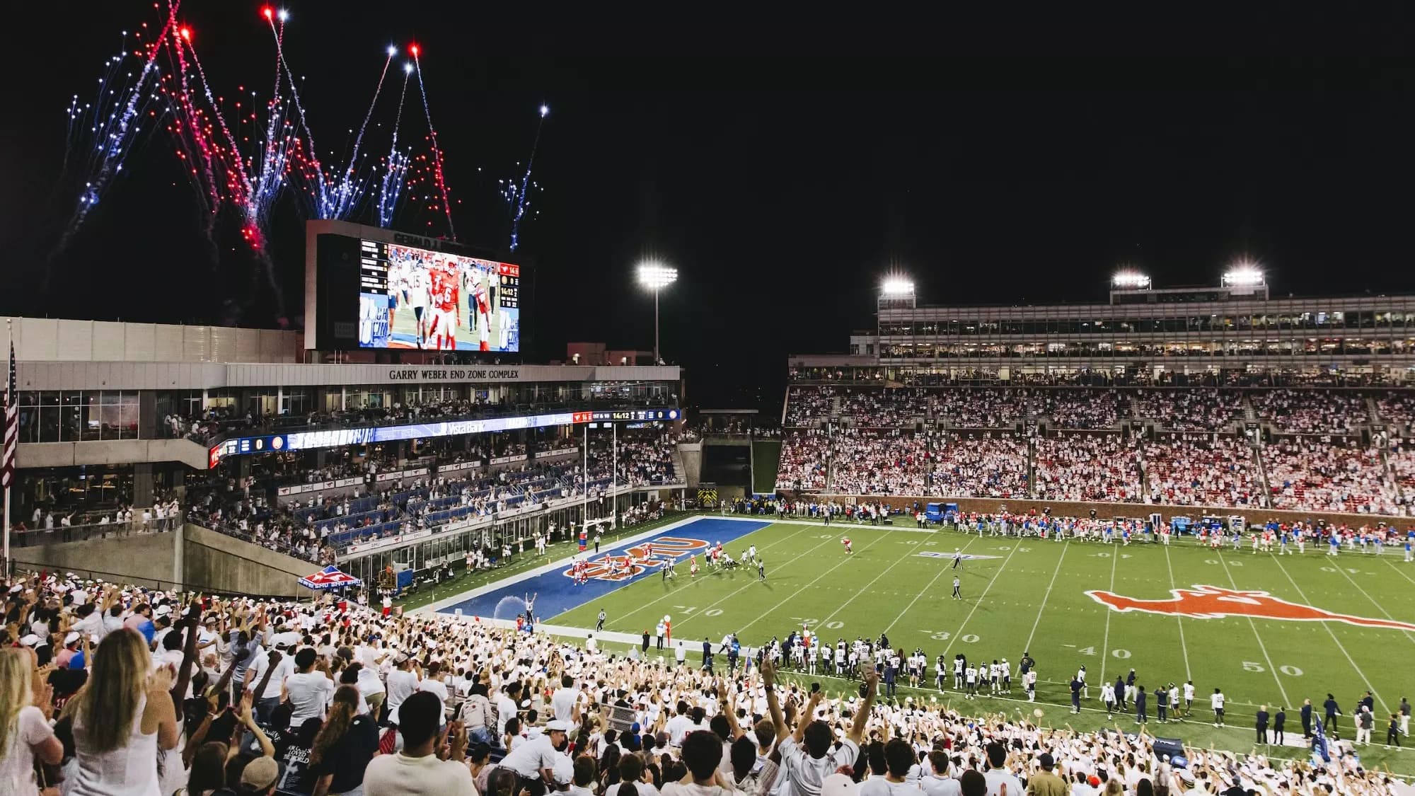 A white-out night crowd packing Gerald J. Ford Stadium as red, white, and blue fireworks burst over the Garry Weber End Zone Complex — the blue SMU end zone and running Mustang at midfield below