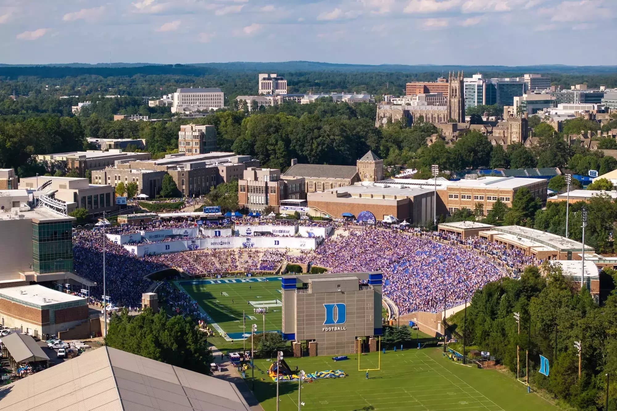 Aerial view of a packed Wallace Wade Stadium on a sunny Saturday — the sunken bowl filled with Duke blue, the Devils Deck banners lining the north end zone, and Duke Chapel's Gothic tower rising from the trees beyond