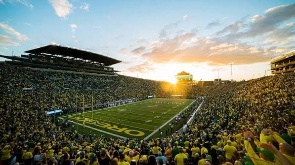 Sunset pouring into Autzen Stadium — a yellow-and-green crowd around the OREGON end zone as the sun drops behind the west rim