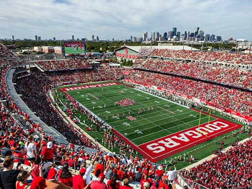 An aerial view of a sold-out Space City Financial Stadium — a packed red-and-white crowd ringing the bowl, COUGARS painted across the end zone, and the downtown Houston skyline rising beyond the stands
