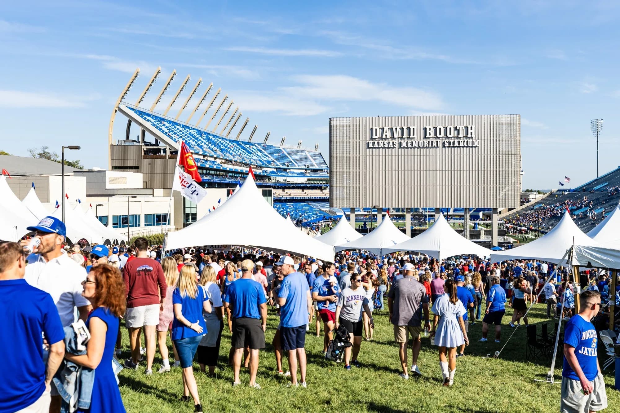 Gameday on the Hill in full swing — white tailgate tents and a blue-clad crowd on the grass above the rebuilt Booth, the new west stands sweeping up behind the DAVID BOOTH KANSAS MEMORIAL STADIUM videoboard under a clear Kansas sky