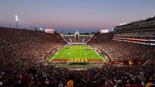 The Los Angeles Memorial Coliseum at dusk — the peristyle arches above a cardinal-and-gold crowd and the USC end zone