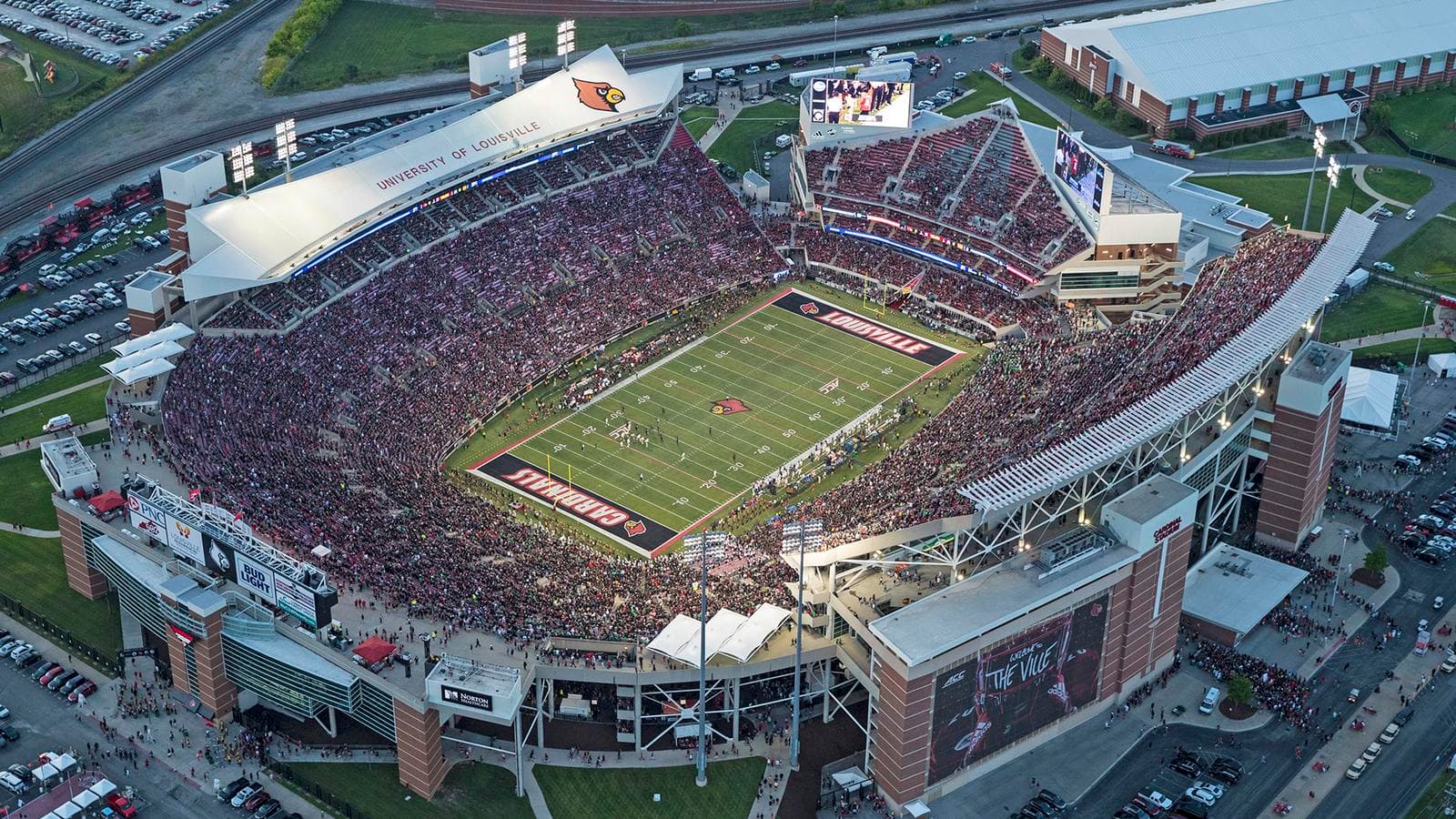 L&N Federal Credit Union Stadium from the air at dusk — a sold-out red-and-black horseshoe under the lights, CARDINALS and LOUISVILLE end zones framing the field, the old rail lines still curving past the north end