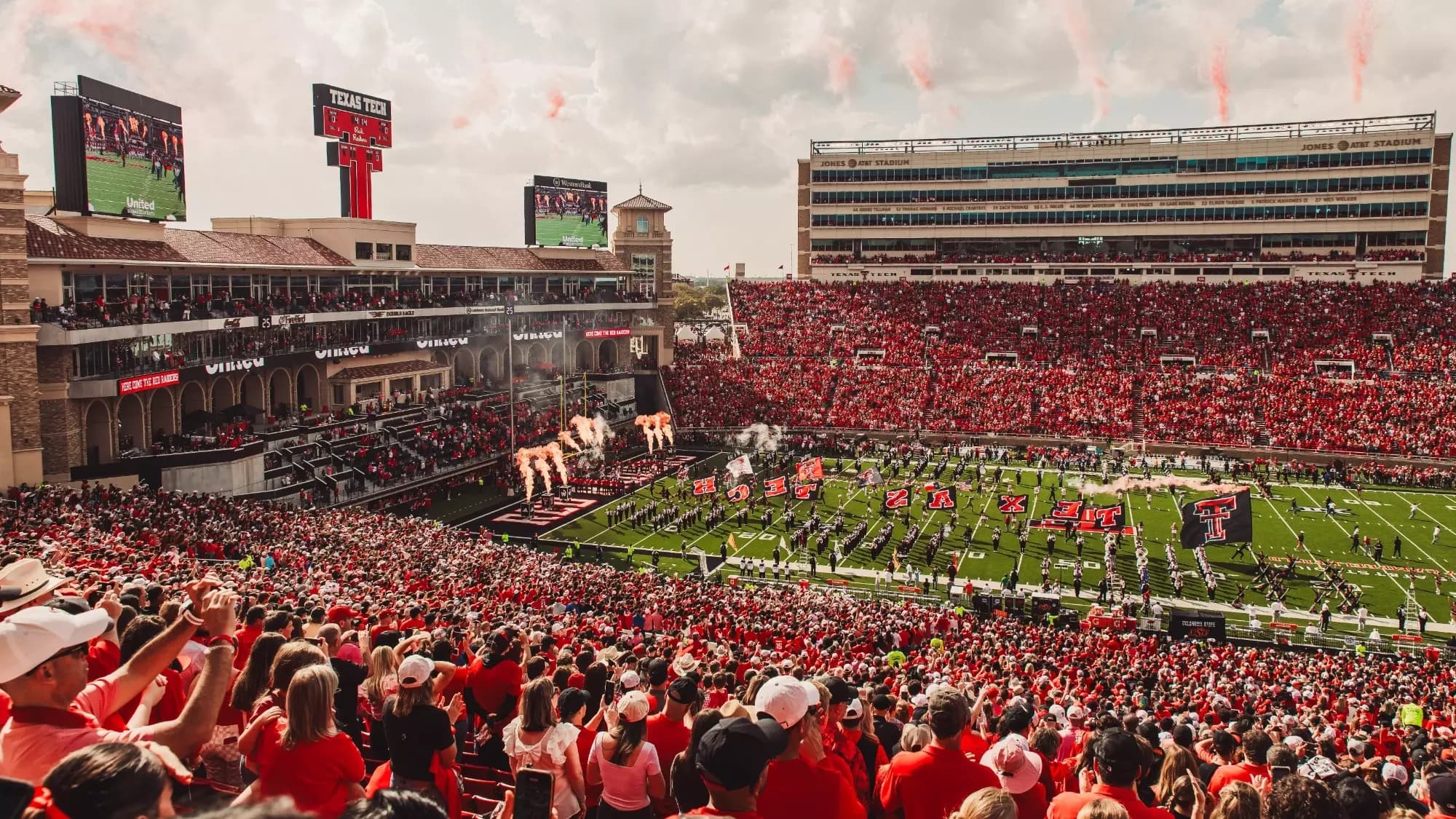 Jones AT&T Stadium at the team entrance — flags spelling TEXAS TECH across the field, pyro flaring, and a sellout crowd in scarlet under a West Texas sky