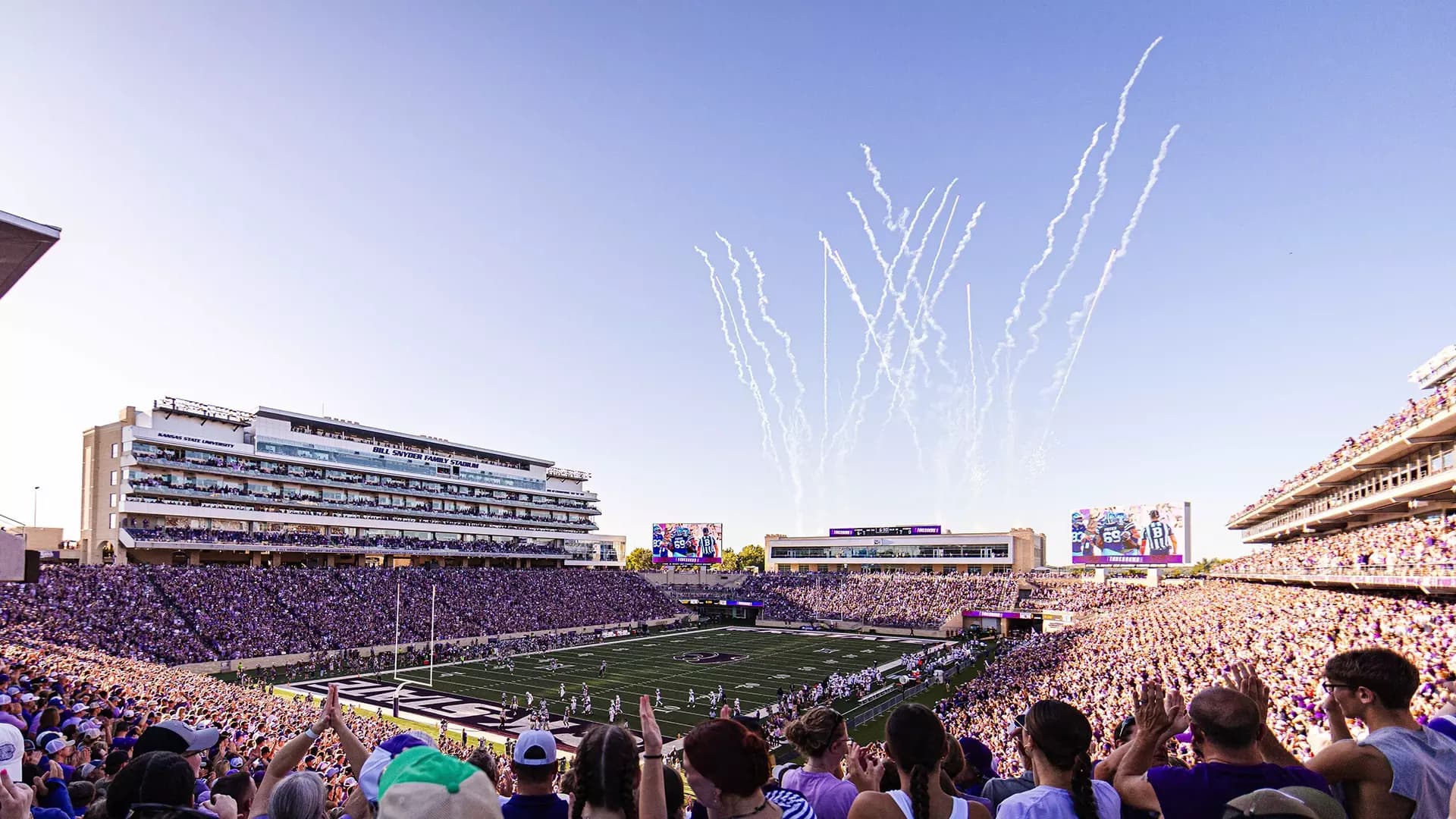 Bill Snyder Family Stadium packed in purple at golden hour — touchdown fireworks streaking over the north videoboards, the KSTATE end zone below, and the press box lettering catching the late Kansas sun
