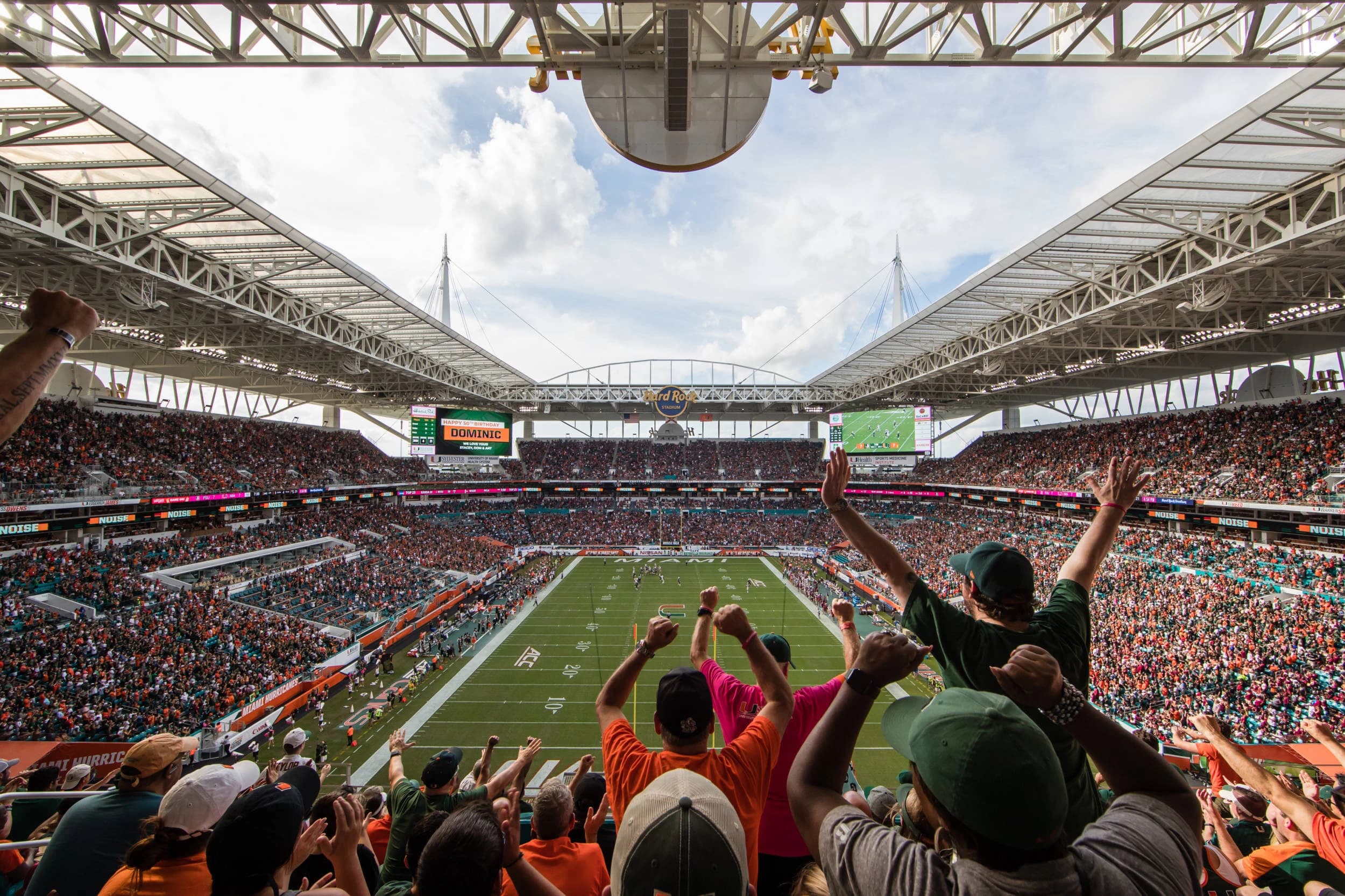 Inside Hard Rock Stadium on a Hurricanes Saturday — fans in orange and green throwing their arms up in the upper deck as the white canopy frames an open square of South Florida sky over the field