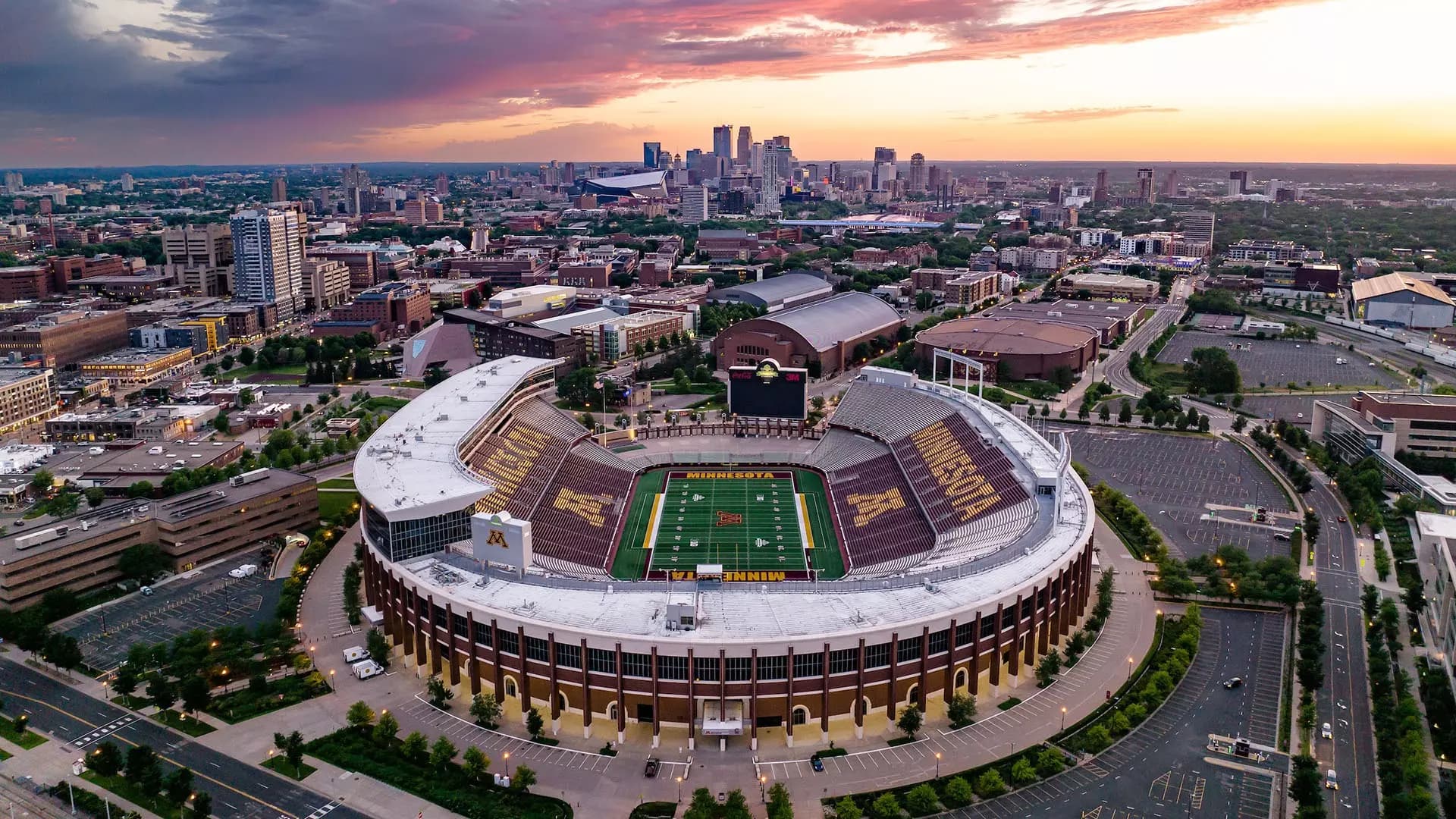 Aerial view of Huntington Bank Stadium at sunset — the maroon-and-gold horseshoe open toward the downtown Minneapolis skyline