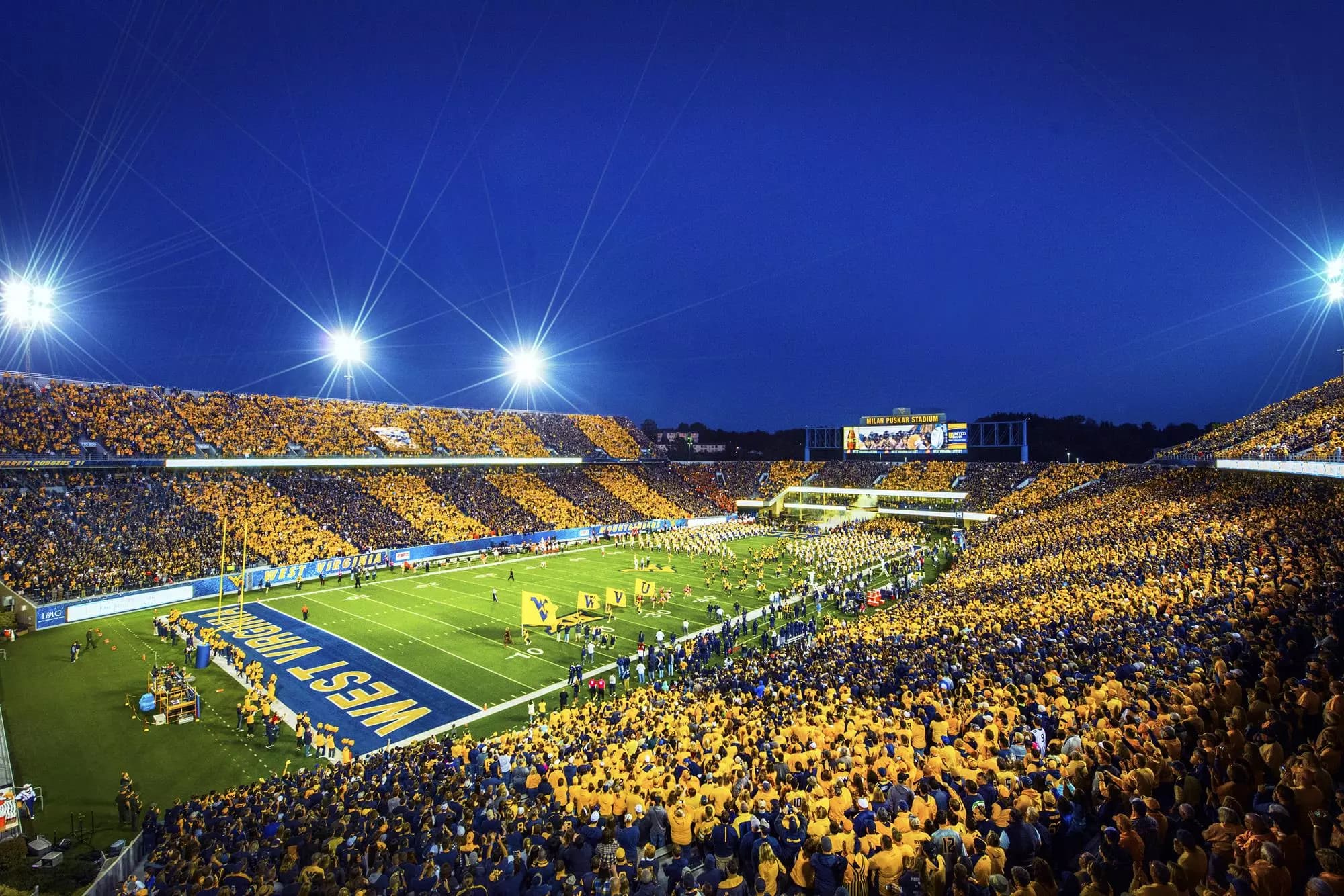 Stripe the Stadium night at Milan Puskar — alternating gold and blue crowd stripes wrapping the full bowl under the lights, the Mountaineers running out past WV flags, MILAN PUSKAR STADIUM on the videoboard against a deep blue sky