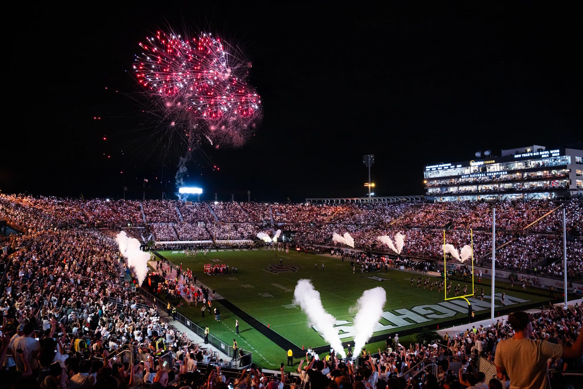 The Bounce House at night — fireworks bursting over a packed bowl lit by thousands of phone lights, smoke jets firing along the KNIGHTS end zone, and the press box banner reading 2017 National Champions