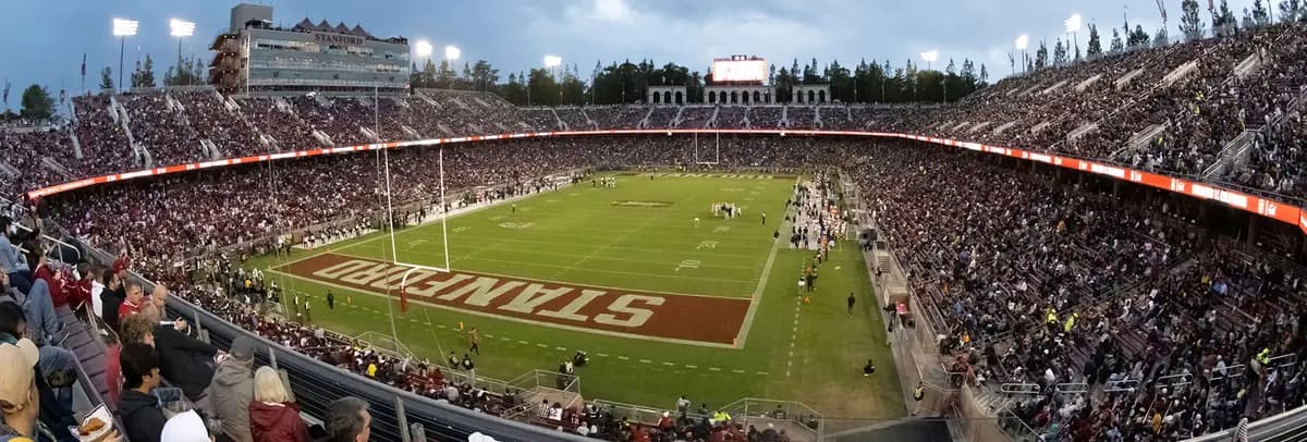Stanford Stadium packed for a night game — the cardinal STANFORD end zone in the foreground, a full bowl of red and white under the lights, the arched colonnade rising over the far rim, and the trees of The Farm against a dusk sky