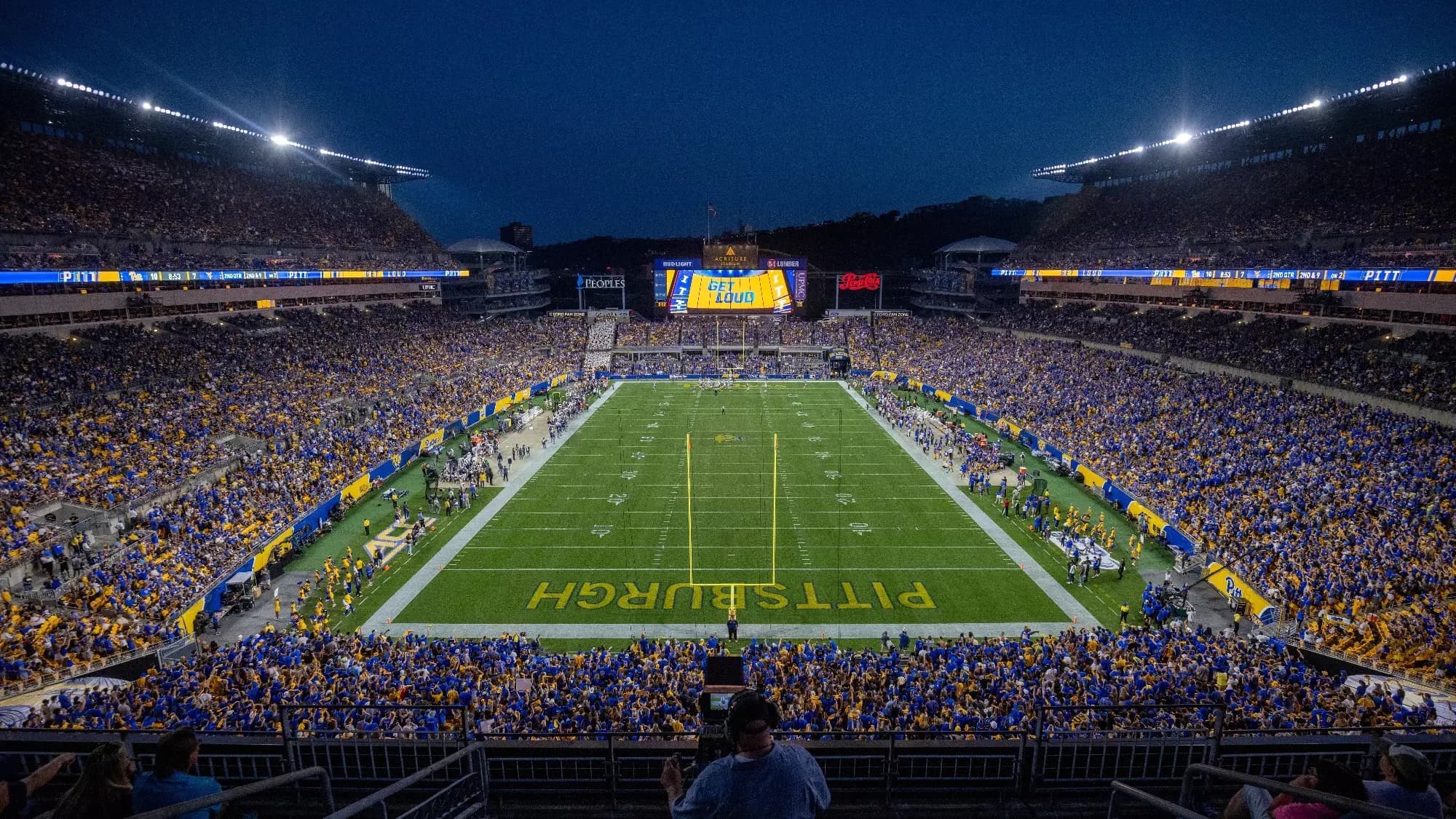 Acrisure Stadium under the lights on a sold-out stripe-out night — the bowl banded in royal blue and gold from the end zone, PITTSBURGH painted across the near end zone and the scoreboard glowing GET LOUD over the river end