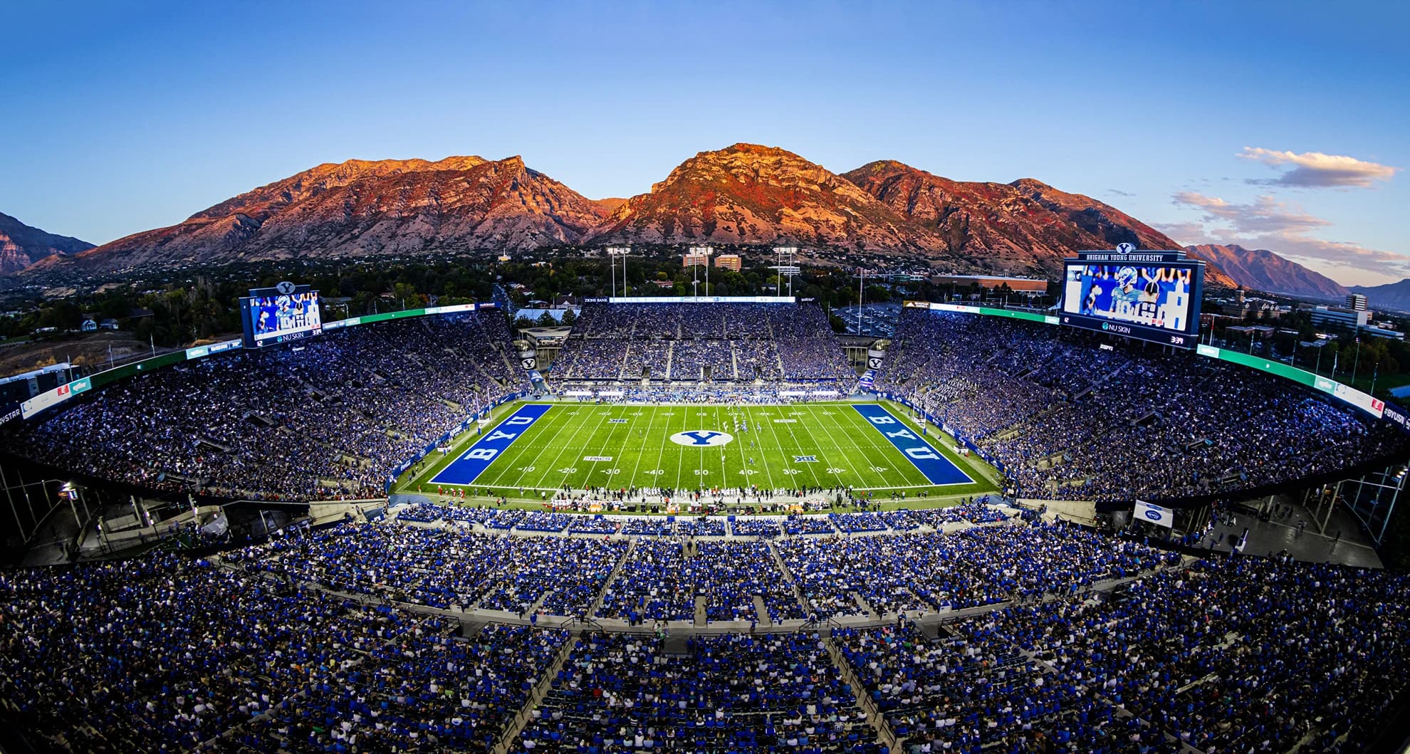 A packed LaVell Edwards Stadium at dusk — a royal-blue sellout crowd, blue BYU end zones framing the white Y at midfield, and the Wasatch Front glowing red with alpenglow straight off the stadium's rim
