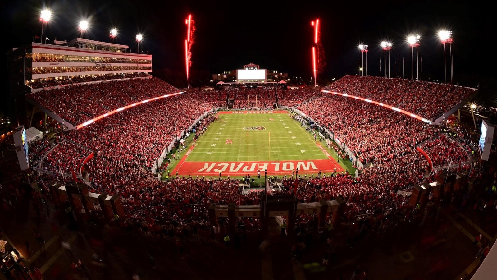 Carter-Finley Stadium at a red-out night game — a packed bowl of Wolfpack red under the lights, red pyro streaking the sky above the press box as the WOLFPACK end zone glows at midfield