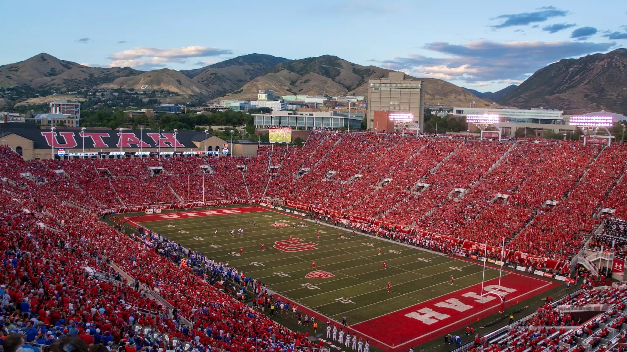 A packed Rice-Eccles Stadium at dusk — a sea of red filling the bowl, the UTAH end wall over the north stands, and the Wasatch foothills behind with the block U visible on the mountainside