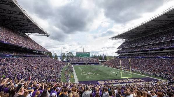 Husky Stadium on a fall Saturday — purple filling both cantilevered grandstands with the HUSKIES end zone below