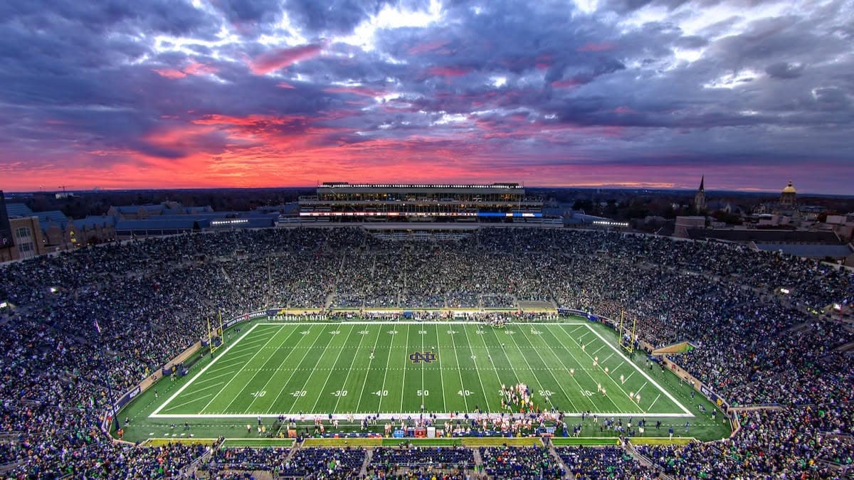 Notre Dame Stadium at sunset — a packed bowl under a dramatic pink-and-purple sky, the Golden Dome visible in the distance beyond the north end zone