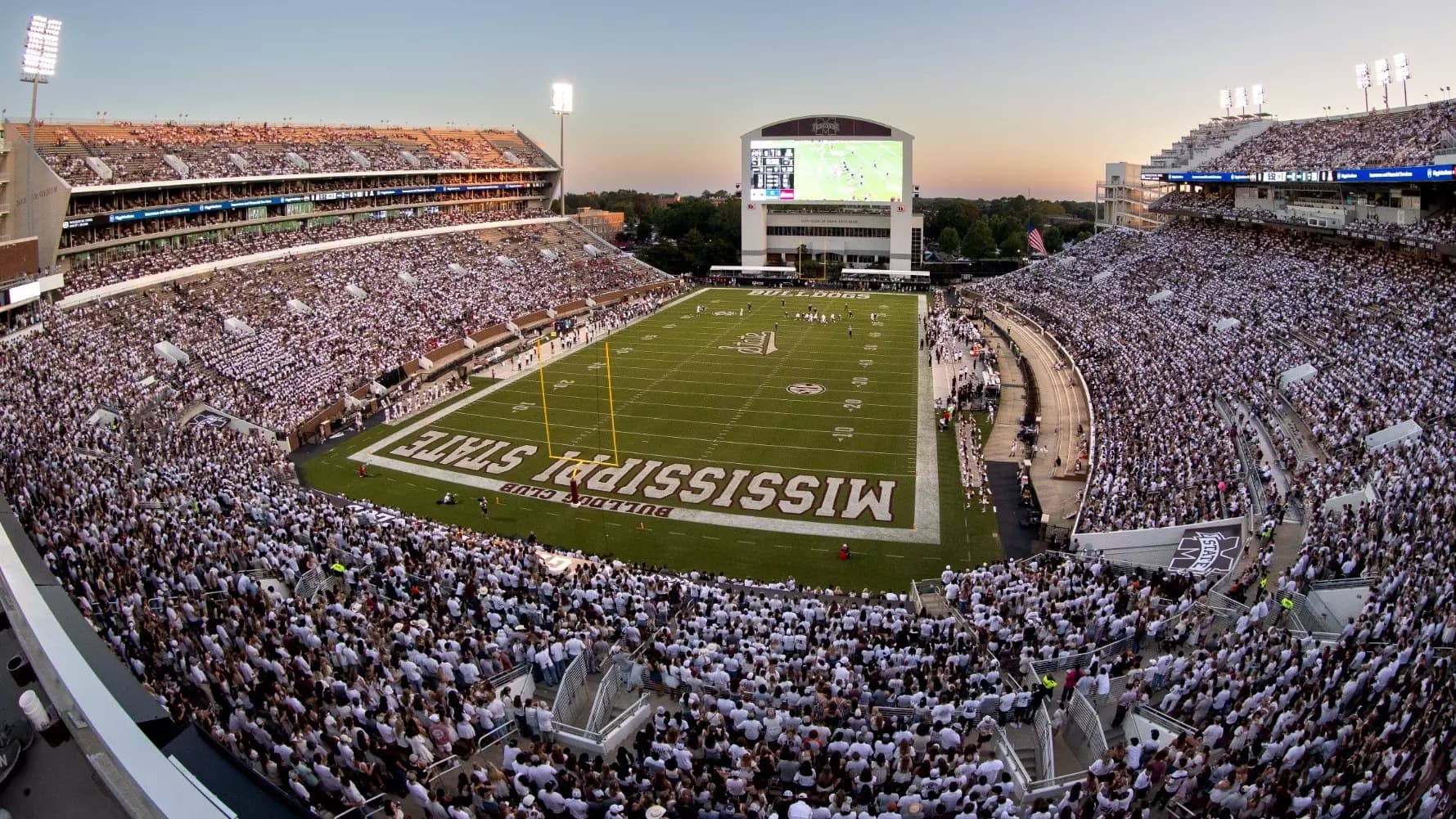 Davis Wade Stadium at Scott Field at dusk during a whiteout — the bowl packed with fans in white, MISSISSIPPI STATE painted across the end zone and the videoboard glowing over a maroon-trimmed field