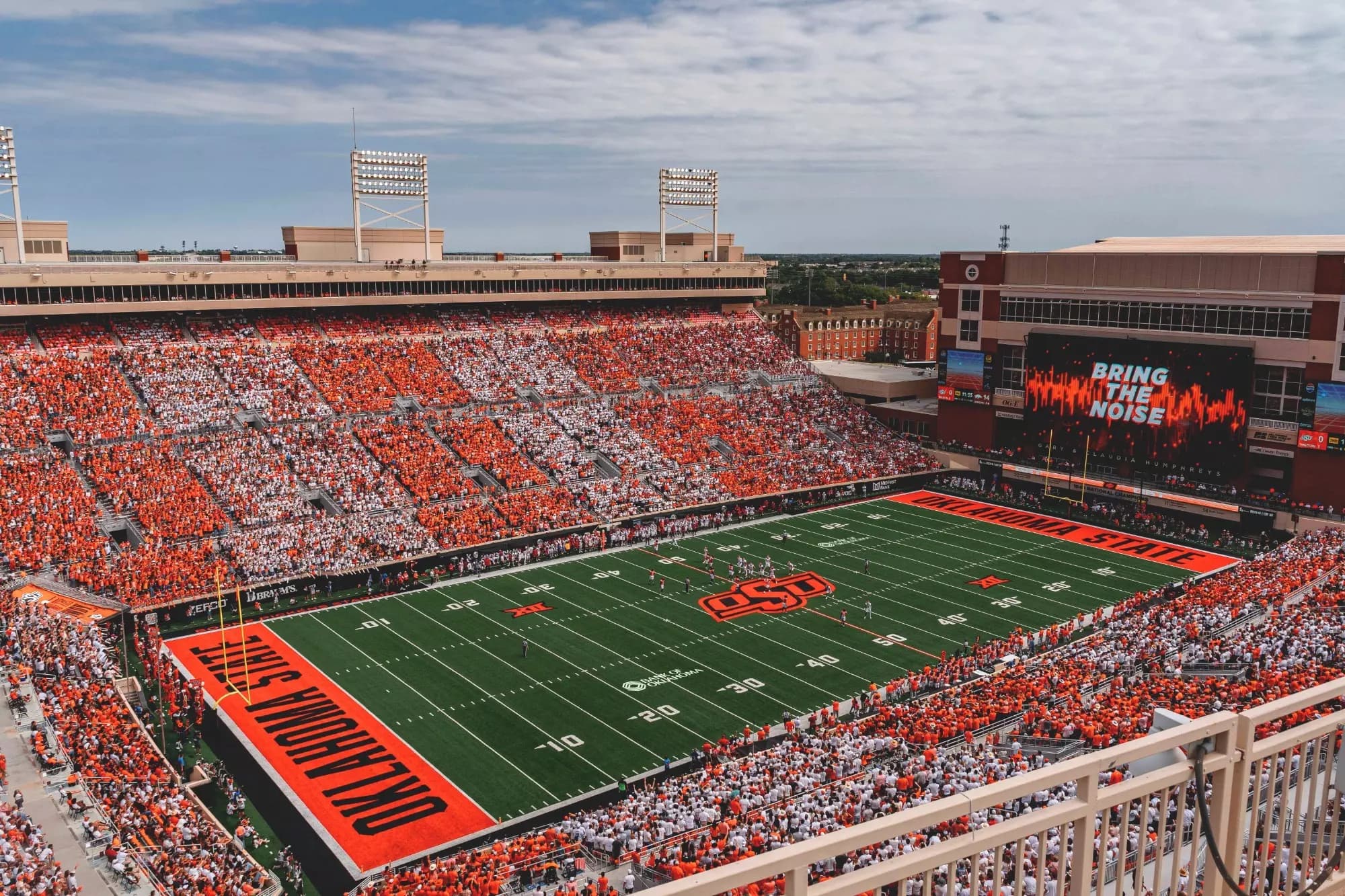 Boone Pickens Stadium striped in alternating orange and white for a sellout — OKLAHOMA STATE end zones framing the sideways field, BRING THE NOISE on the videoboard, and the Oklahoma prairie stretching flat past the campus brick beyond the rim