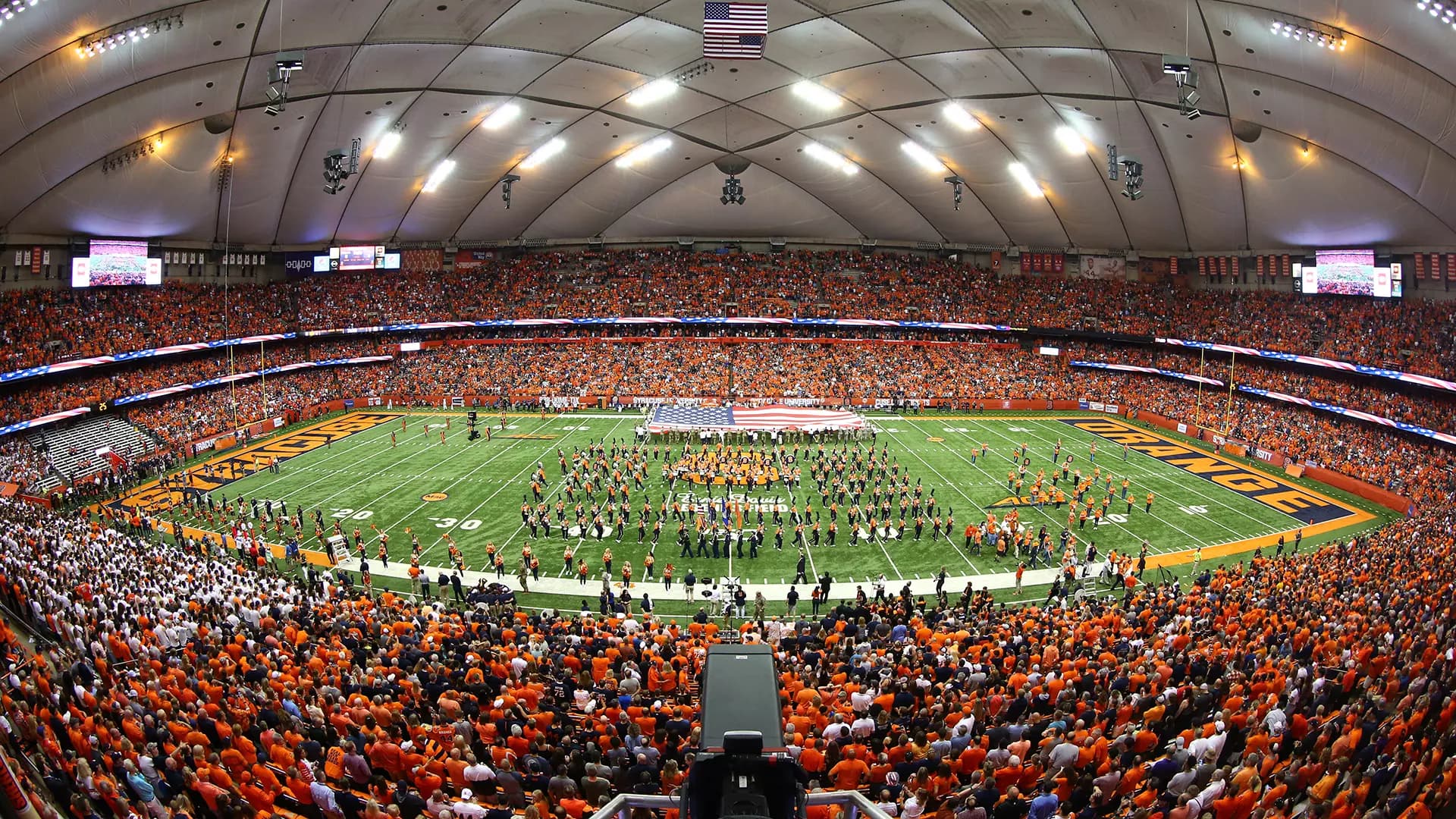 A sold-out JMA Wireless Dome from the upper bowl — the stands a solid wall of orange under the white roof, the marching band spread across the field around a giant American flag with ORANGE painted in the end zone