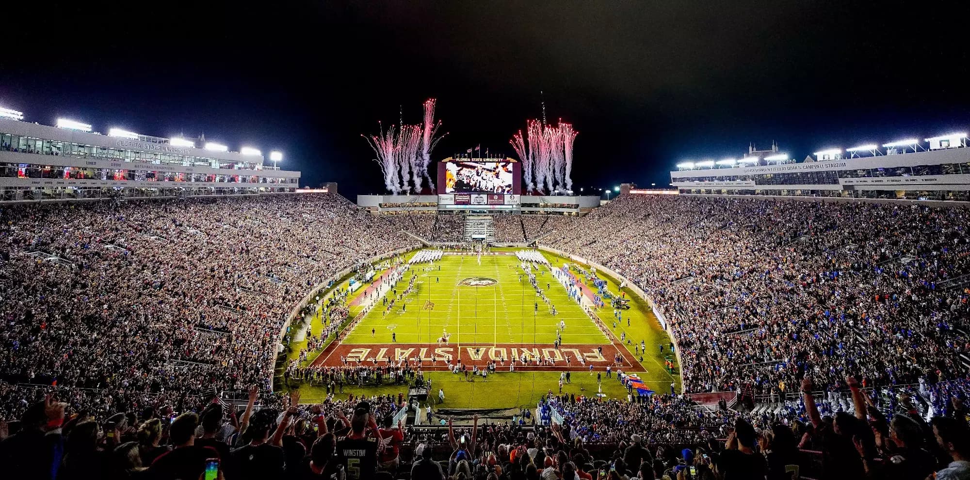 Doak Campbell Stadium at night — fireworks bursting over the videoboard, a full garnet-and-gold house, and Osceola and Renegade on the grass near the FLORIDA STATE end zone