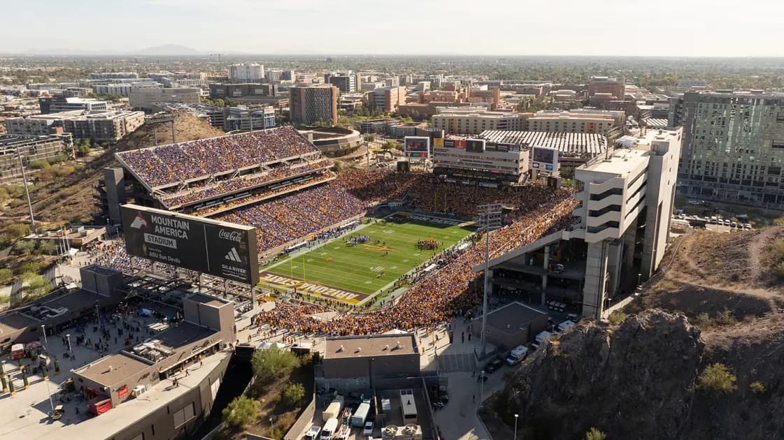 Aerial view of a packed Mountain America Stadium on game day — the bowl wedged against Hayden Butte's rock face, a gold-and-maroon crowd, and downtown Tempe spreading beyond the stands