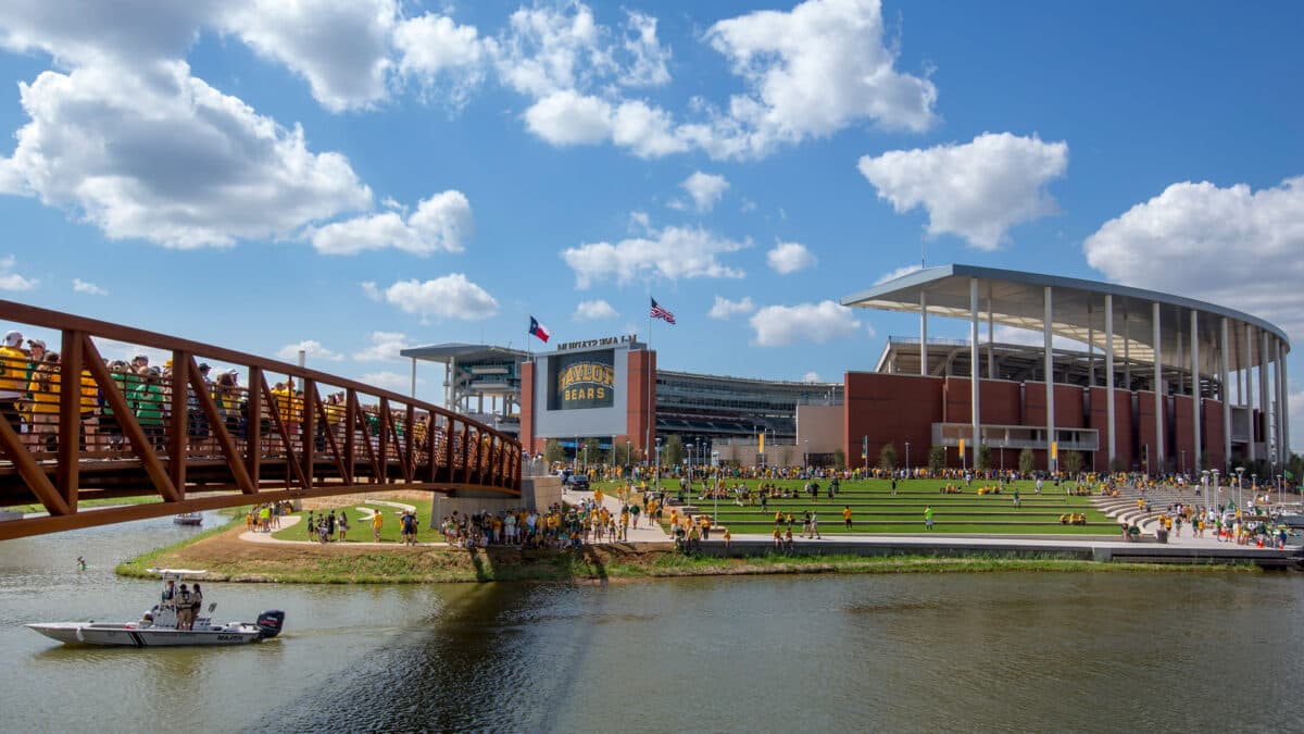 McLane Stadium from across the Brazos River on game day — fans crossing the Umphrey Pedestrian Bridge, green and gold filling the riverside steps, a boat idling below