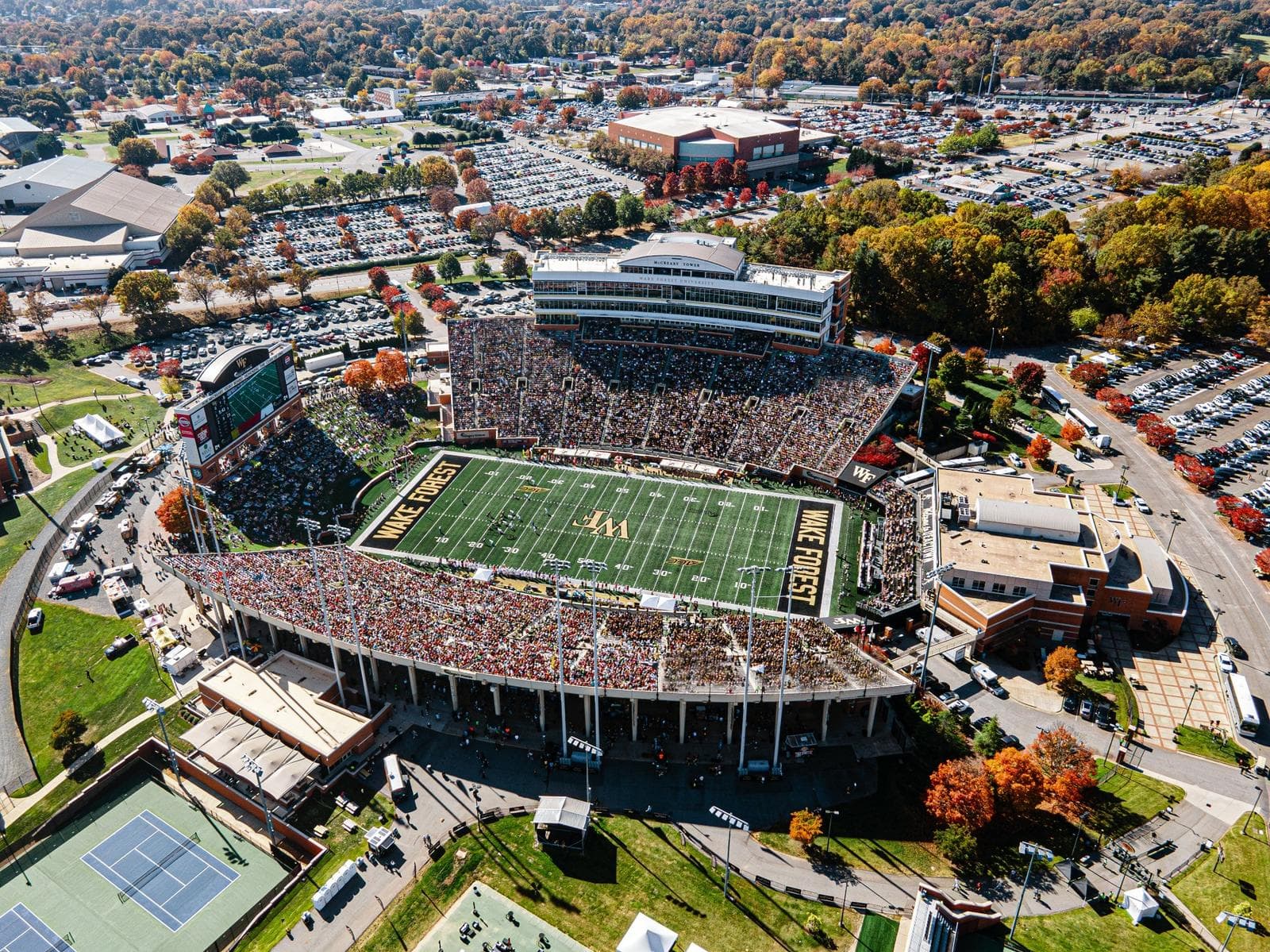 Allegacy Stadium sold out on a peak-fall Saturday from the air — WAKE FOREST in gold across both end zones, McCreary Tower over the press box, and autumn color ringing the full tailgate lots