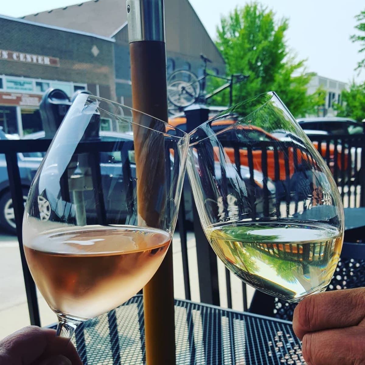 Two glasses of rosé and white wine clinking together on Della Viti's outdoor patio table, downtown Ames storefronts in the background