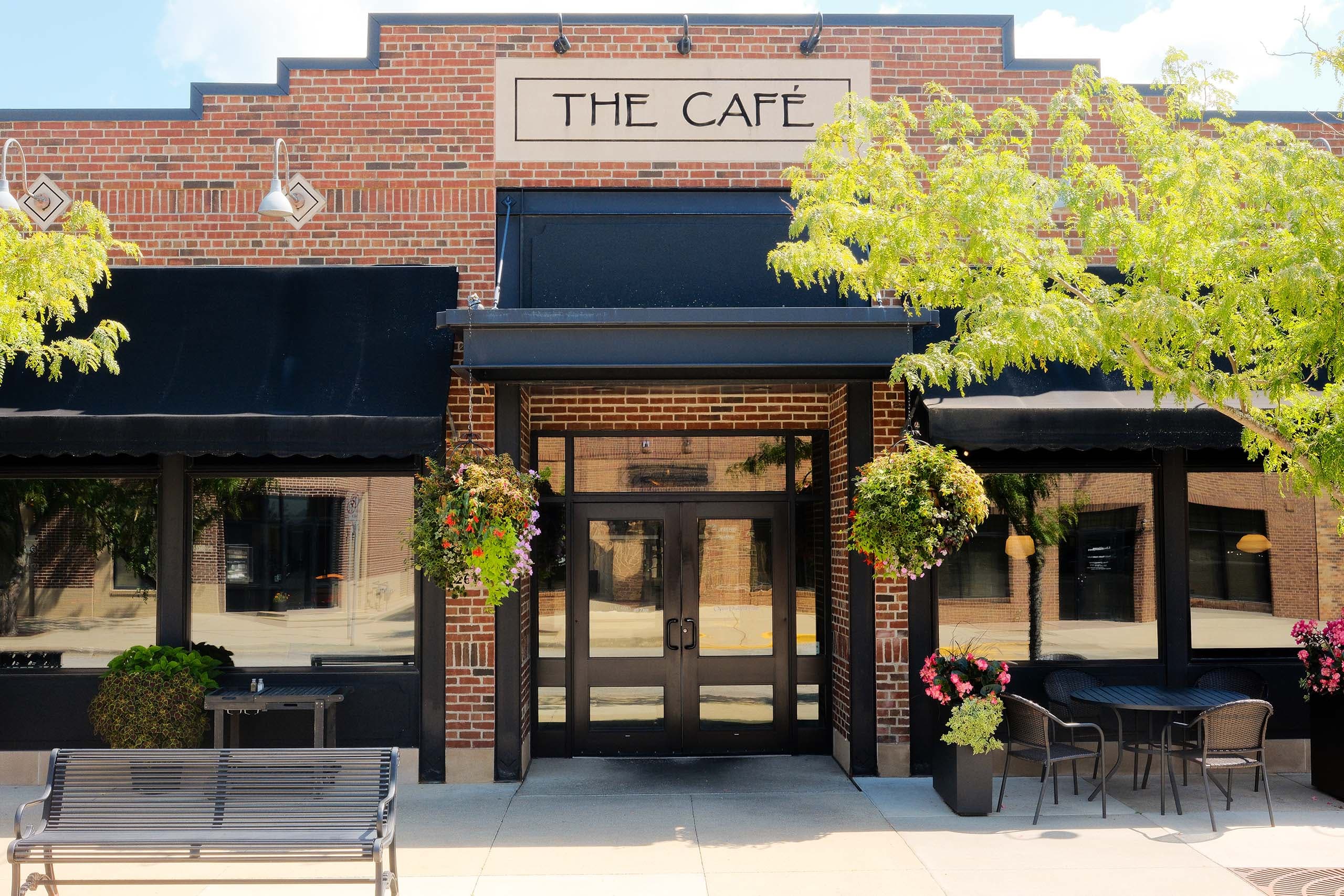 The Café's brick storefront entrance in downtown Ames, with black awnings, hanging flower baskets, and outdoor patio seating