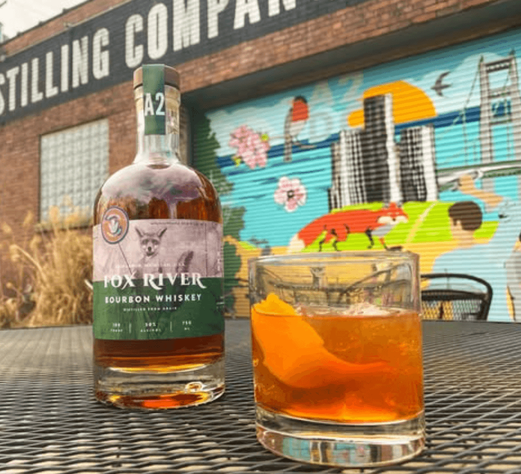 A bottle of Fox River Bourbon Whiskey next to an Old Fashioned on an outdoor table at Ann Arbor Distilling Company, with the distillery's mural in the background