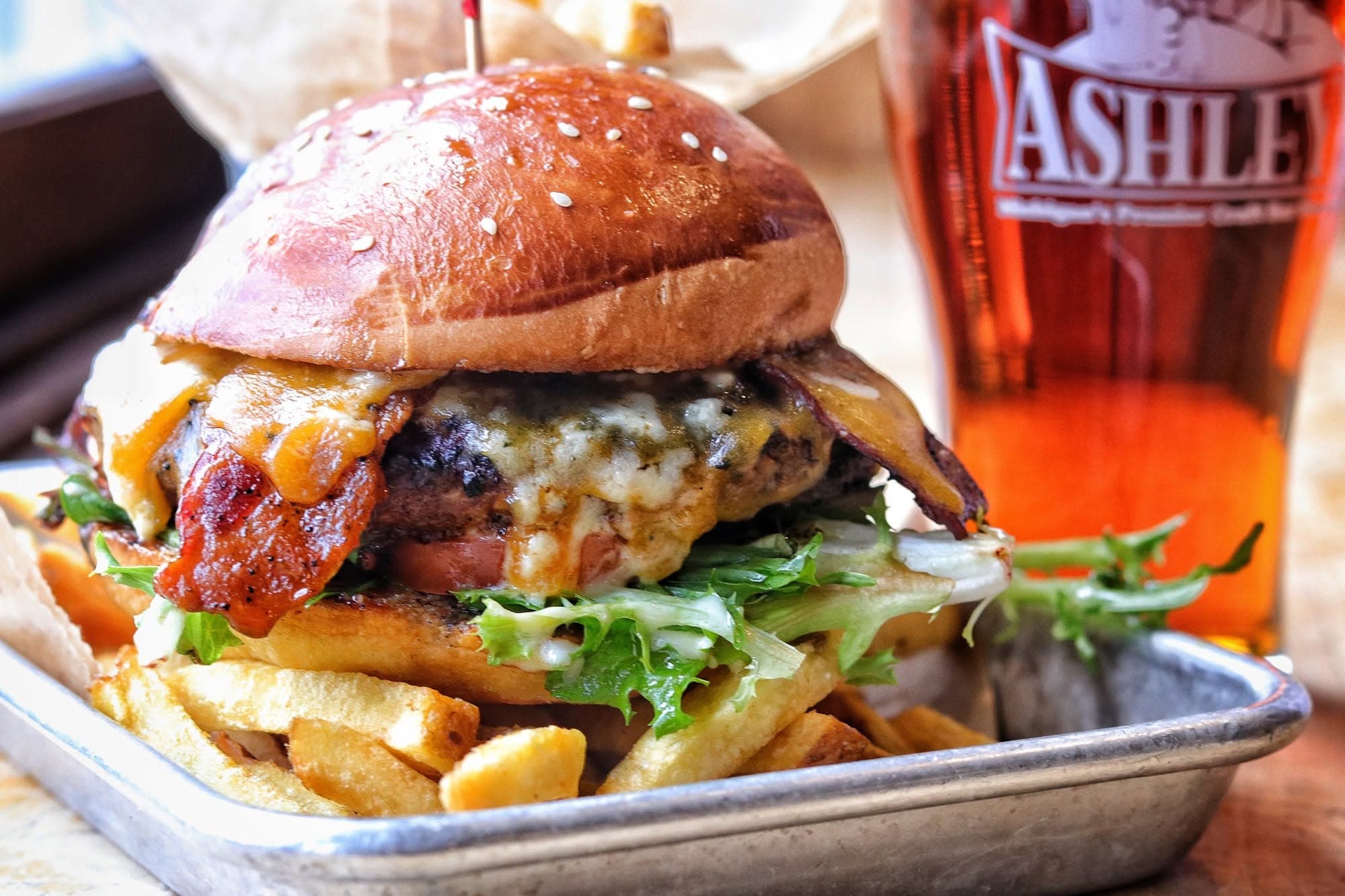 A loaded bacon cheeseburger with fries in a metal basket next to a pint of beer in an Ashley's branded glass