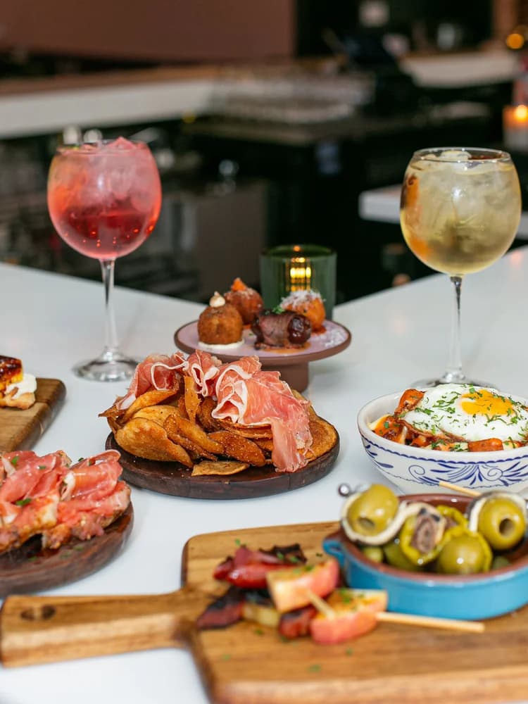 A spread of Aventura tapas — jamón and crisps, a poached-egg toast, marinated olives, and cocktails