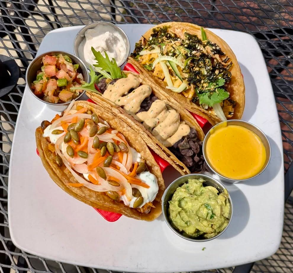 A plate of four loaded plant-based tacos with toppings like pepitas, pickled onion, and kale slaw, served with guacamole, salsa, and queso at Detroit Street Filling Station