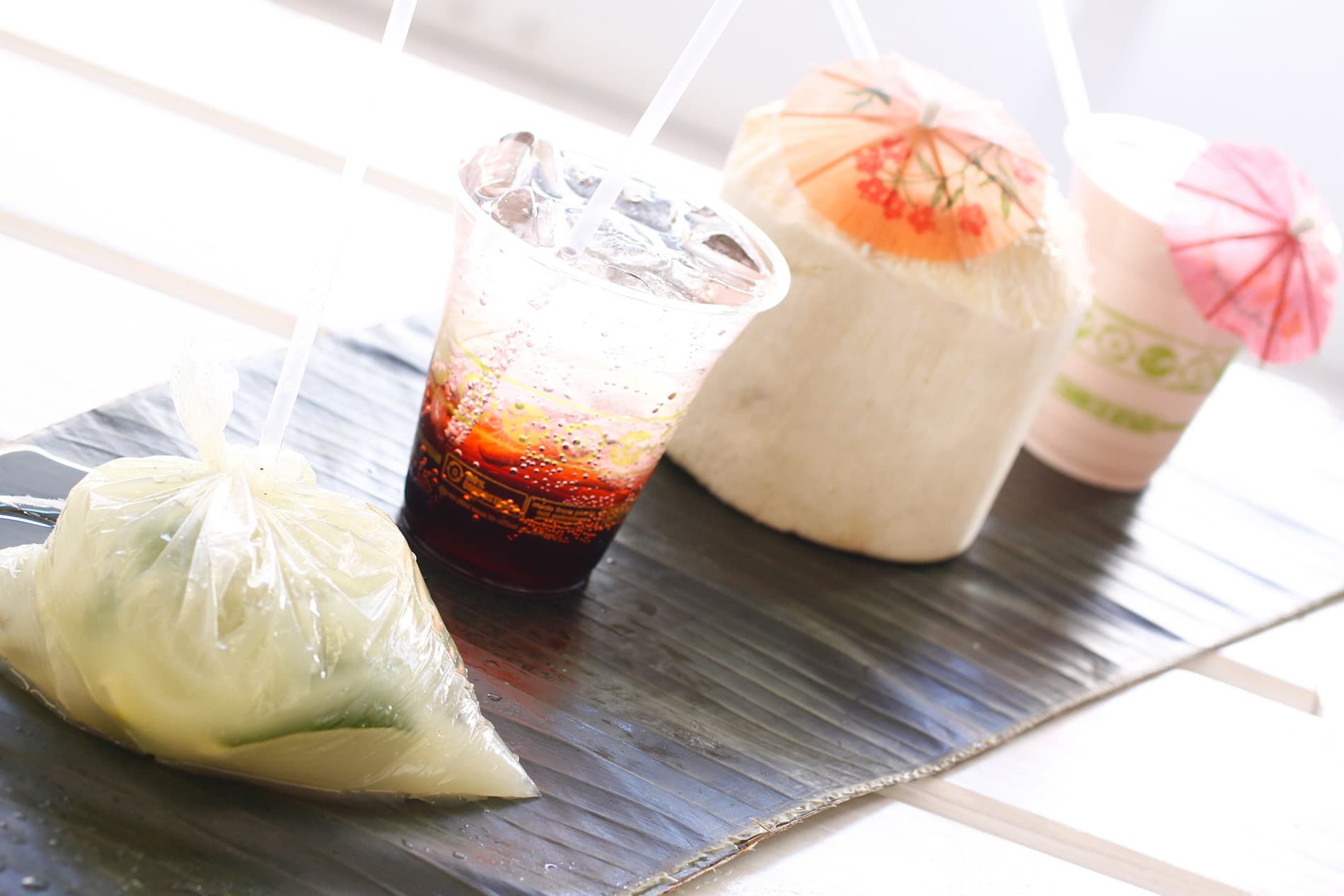 A young coconut with a paper umbrella, a bagged batido, and iced tropical drinks served over banana leaf at Frita Batidos