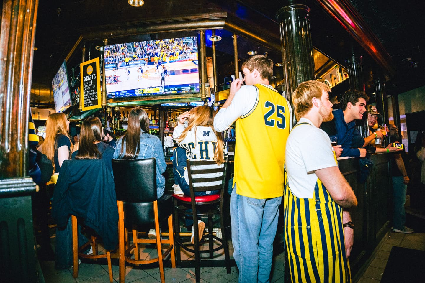 A packed bar at Good Time Charley's with fans in maize and blue watching a Michigan basketball game on the TV overhead