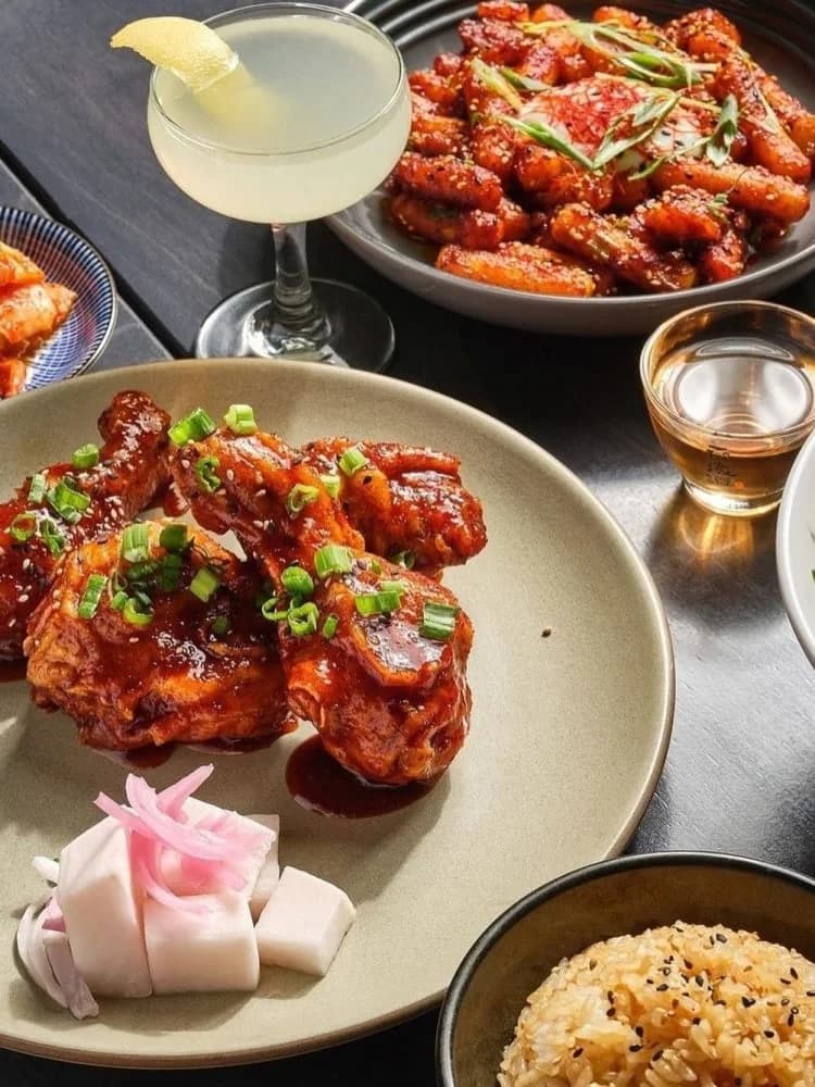 A spread of Korean fried chicken wings with scallions, tteokbokki, pickled radish, a cocktail, and sesame rice at Miss Kim
