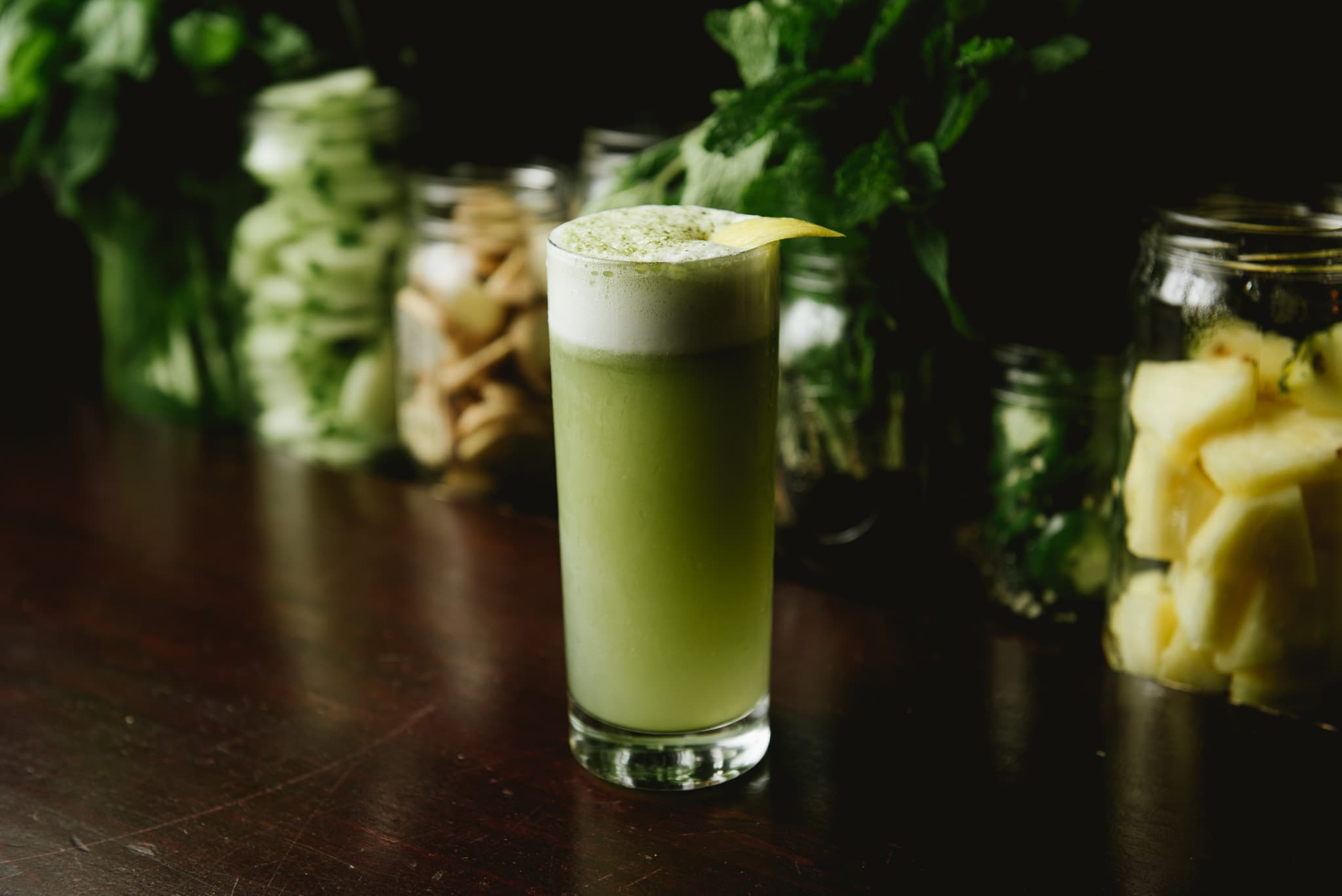 A frothy green cocktail with a lemon twist garnish at The Last Word, backed by jars of fresh herbs and pineapple
