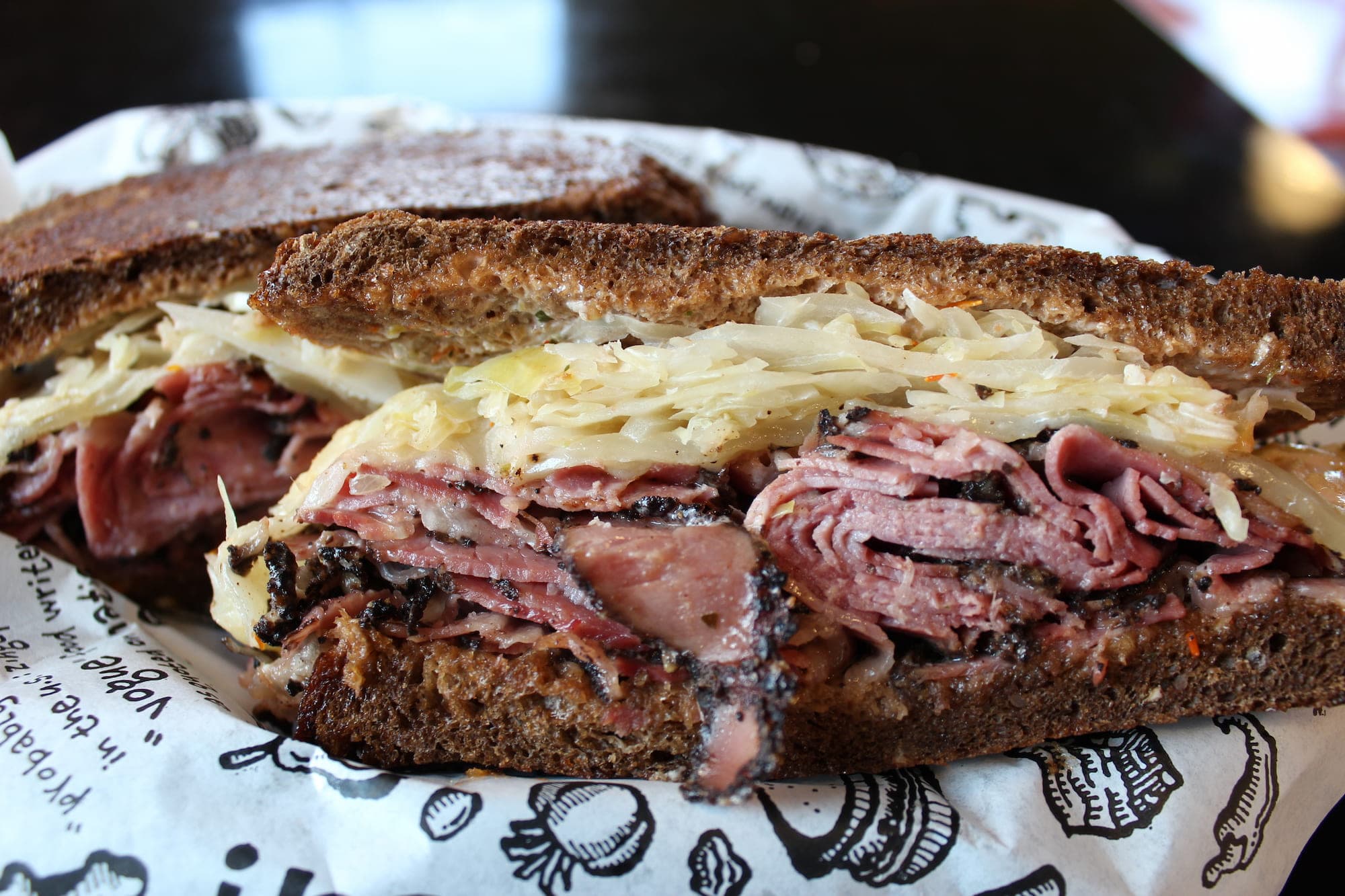 A stacked pastrami and corned beef sandwich on rye with sauerkraut, cut in half, in Zingerman's branded deli paper
