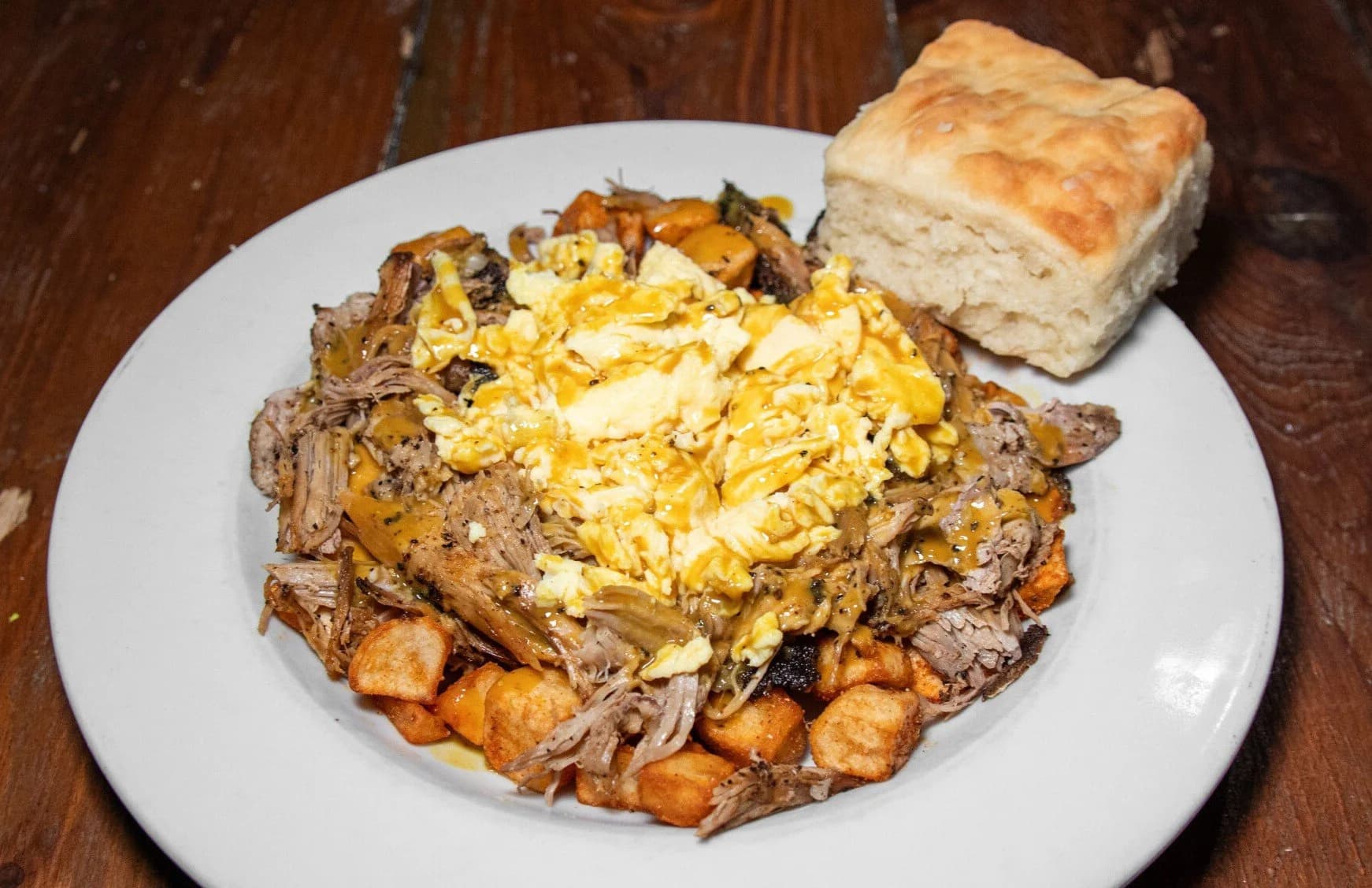 A loaded breakfast plate at Mama's Boy — pulled pork, scrambled eggs, and potatoes drizzled with sauce, next to a tall biscuit