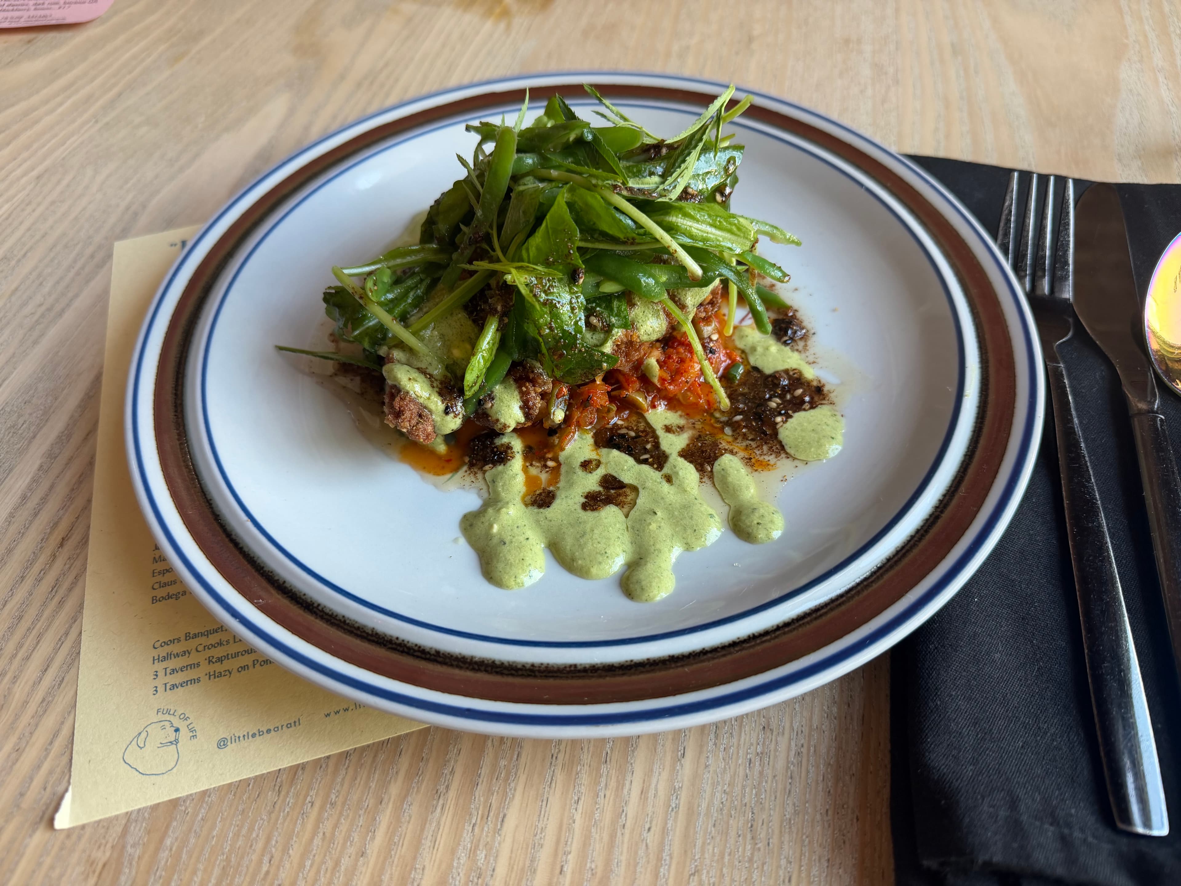 A composed dish of crispy meat topped with herbs and pea shoots, drizzled with green herb sauce, on a striped plate at Little Bear
